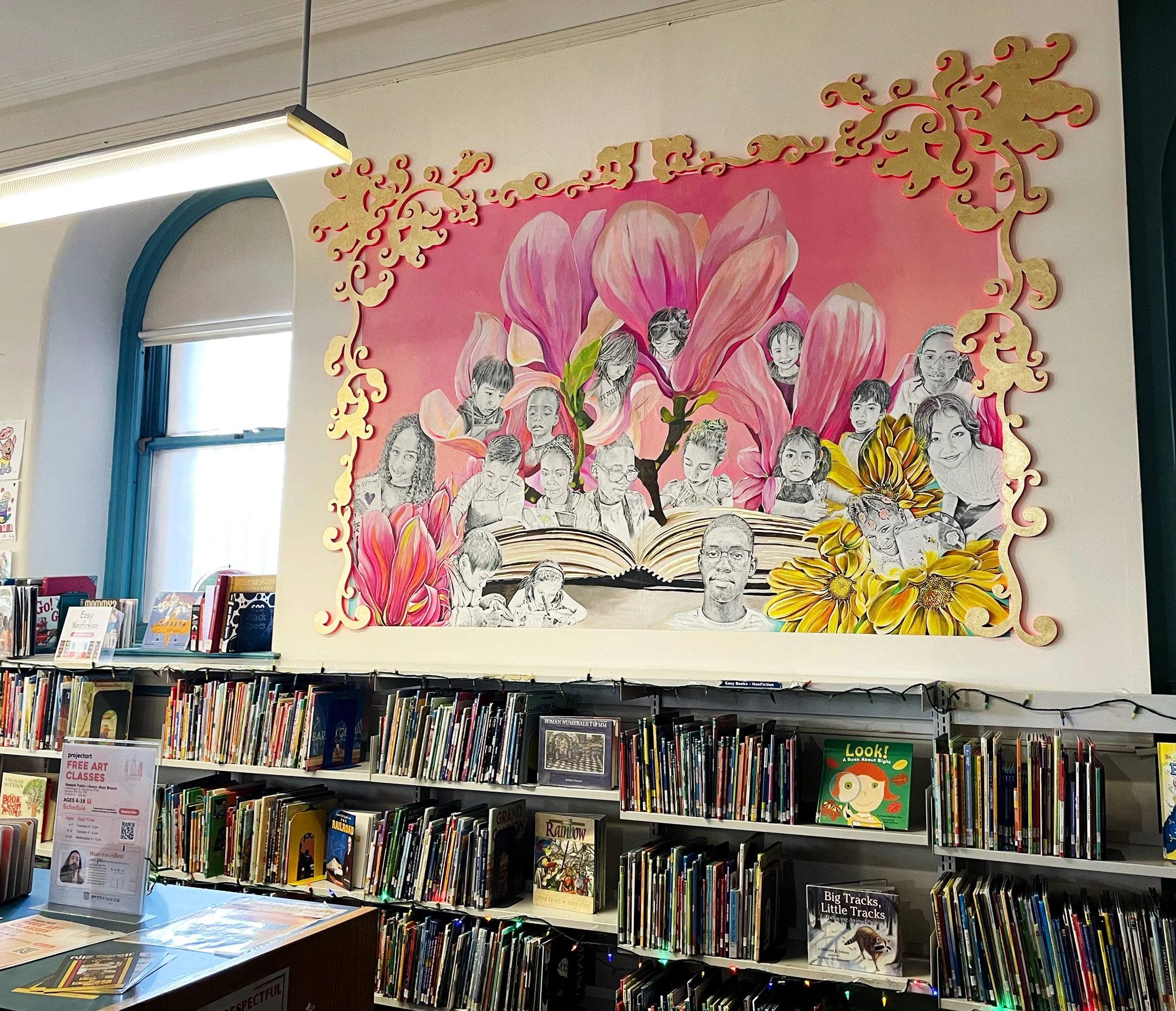 A mural in a library depicting children and adults with large pink and yellow floral elements, framed by a decorative gold border.