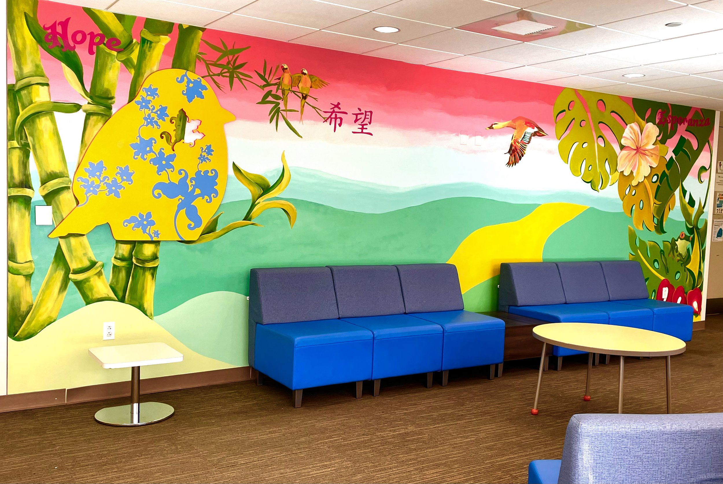 Colorful mural in a waiting area featuring bamboo, birds, a monkey with floral patterns, lush green hills, and tropical foliage, with blue seating and small tables.