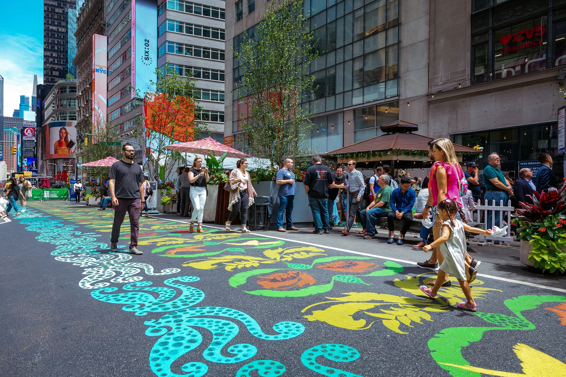 A busy city street with colorful street art on the pavement, people walking, sitting, and standing in front of storefronts and outdoor seating areas, with tall buildings and digital billboards in the background.