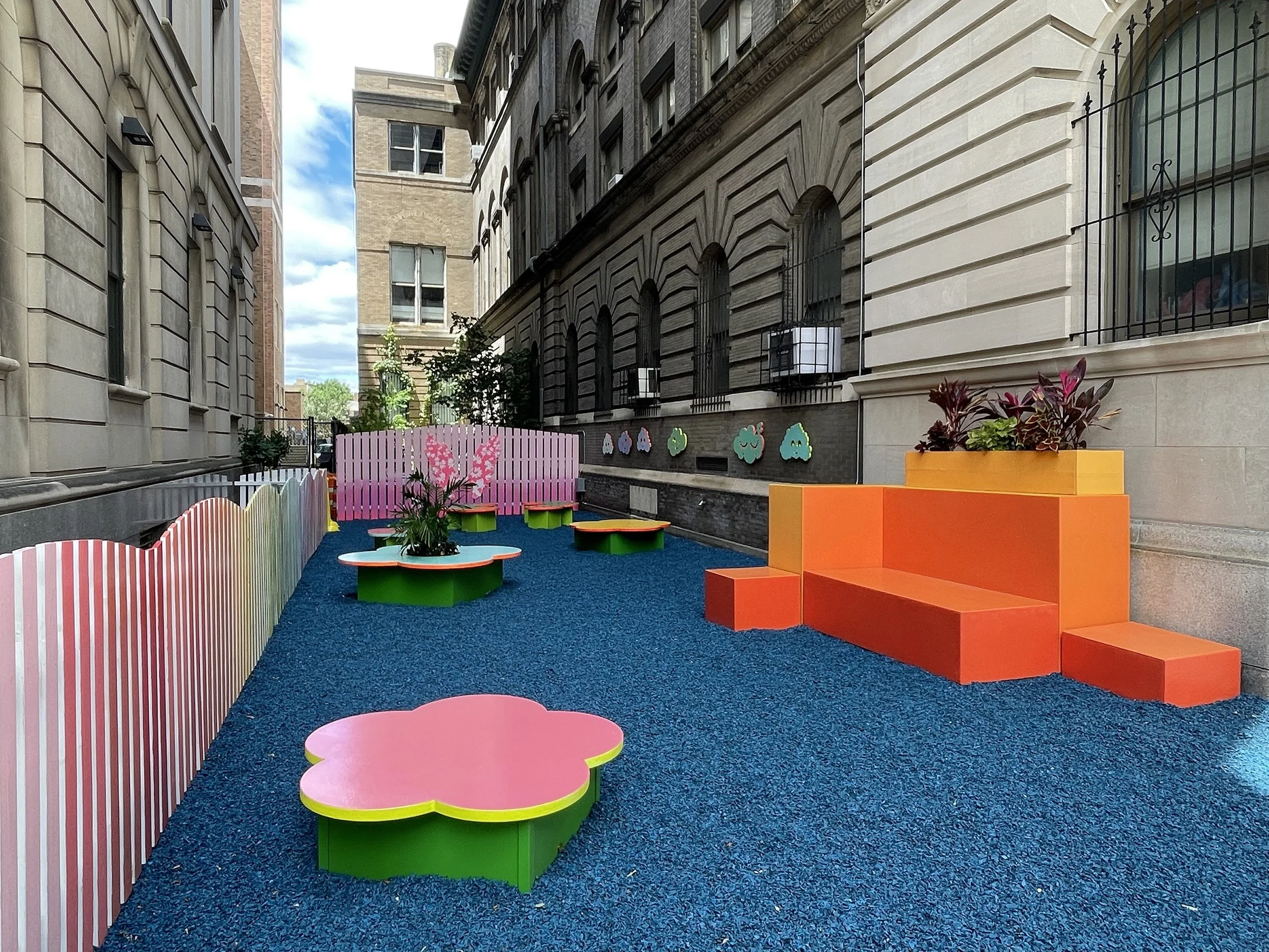 Colorful outdoor urban playground with pink, green, orange, and yellow furniture and a blue rubber ground, enclosed by a rainbow-colored picket fence, with decorative clouds on the building walls and plants on the furniture.