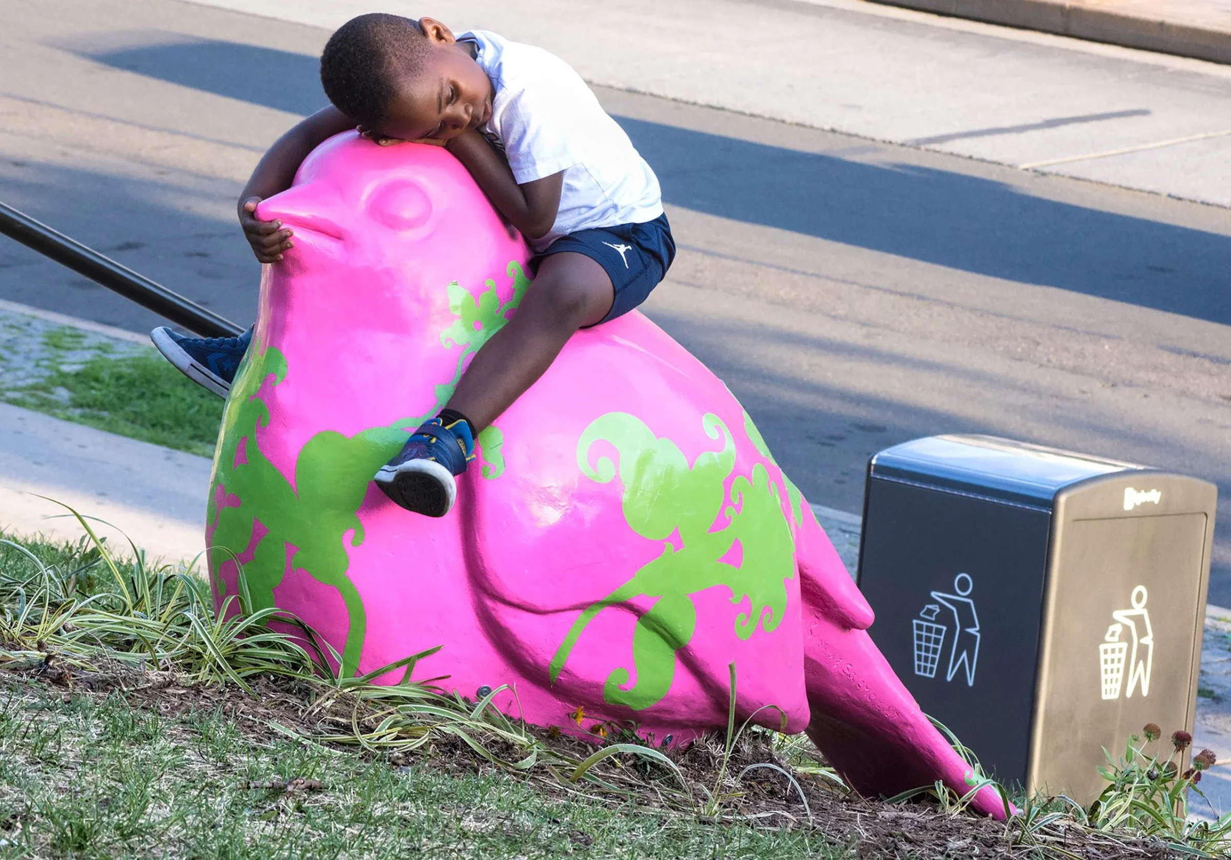 A young boy in a white shirt and navy shorts hugging and sitting on a large pink cow-shaped sculpture with green decorative patterns, located outdoors by a sidewalk. A recycling bin is nearby.