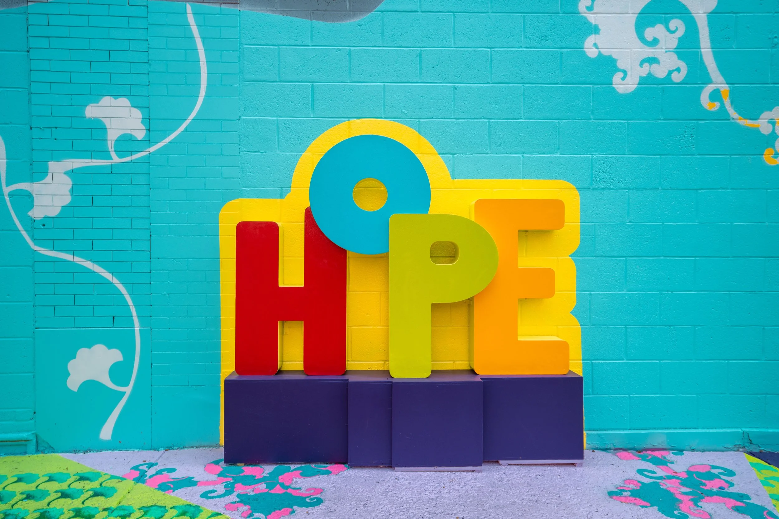 Colorful alphabet letters spelling 'Hope' in front of a bright turquoise wall with decorative white and gold swirls.