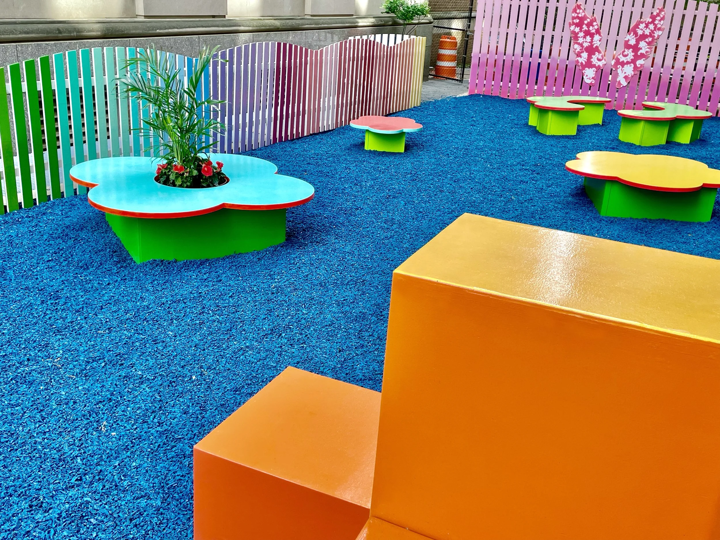 Colorful outdoor children’s play area with flower-shaped seating, a painted bright blue ground, a rainbow-colored fence, and pink wall with butterfly designs.
