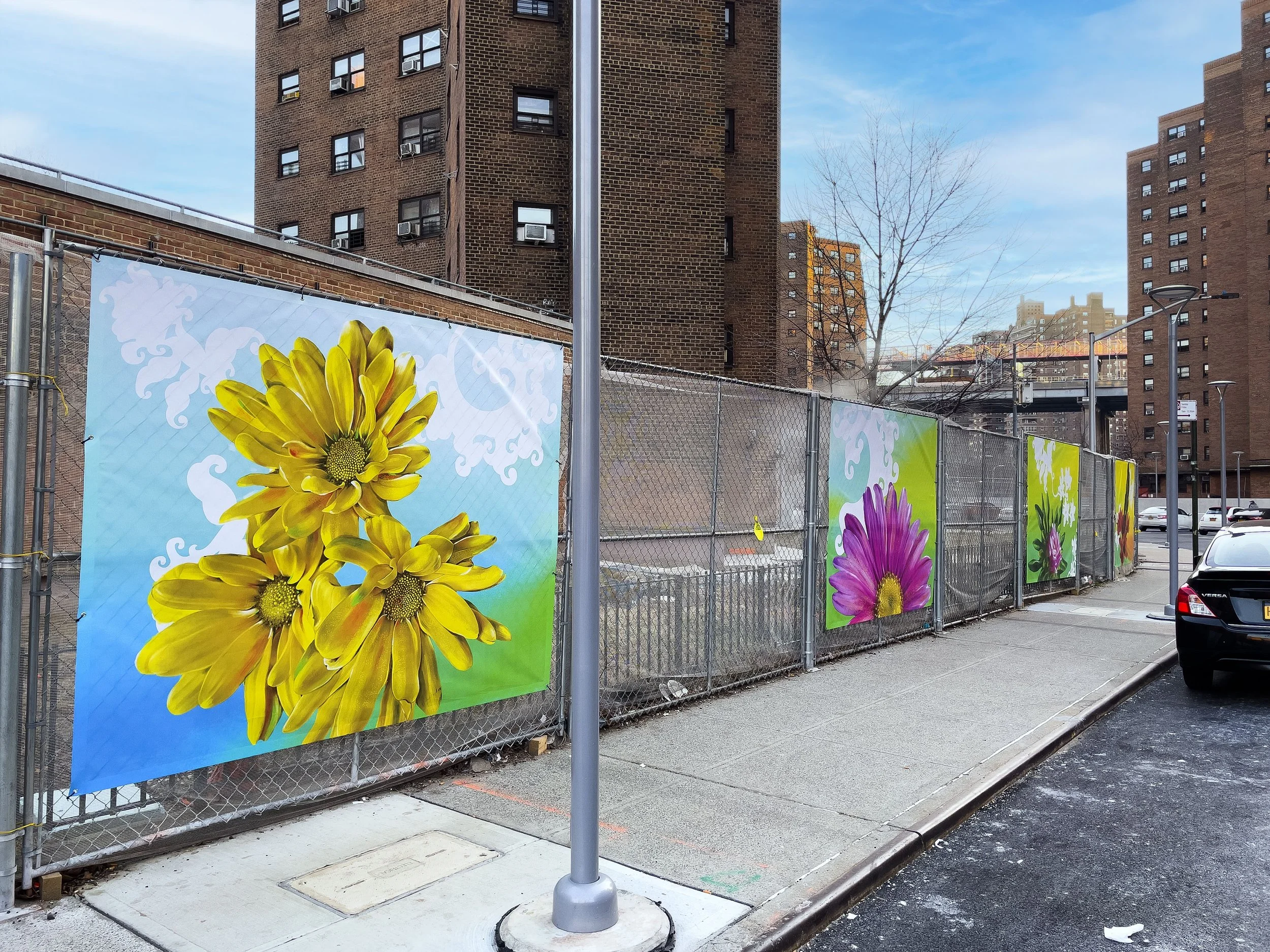Colorful flower posters on a chain-link fence on an urban sidewalk with brick apartment buildings in the background