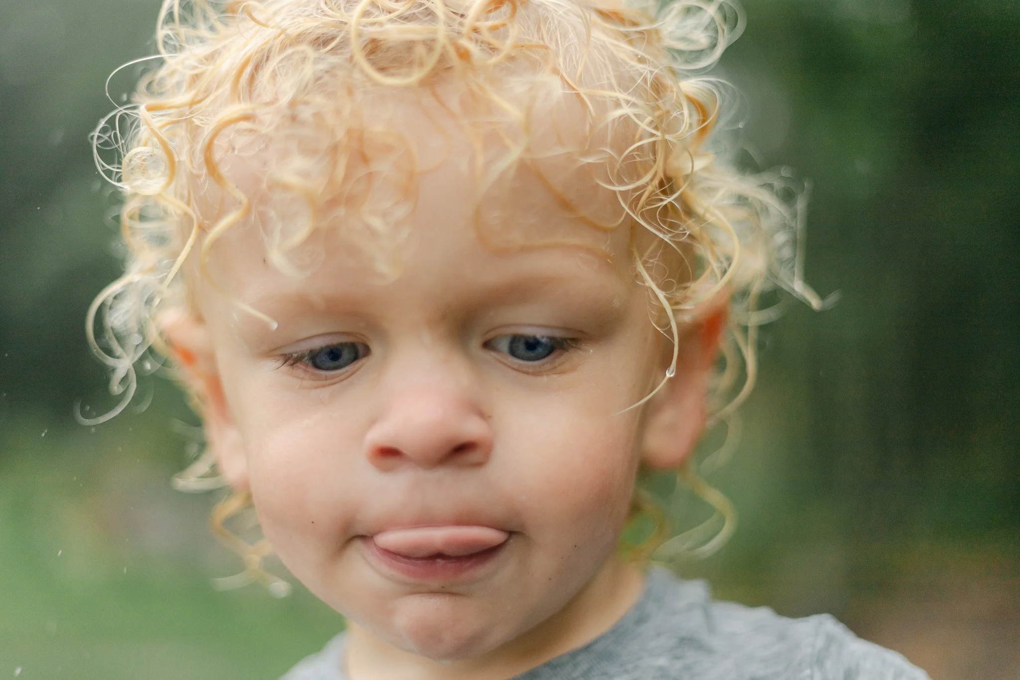 A young boy with curly blonde hair, blue eyes, and light skin sticking out his tongue, outdoors with a green background. Photographer in Franklin, Wisconsin.