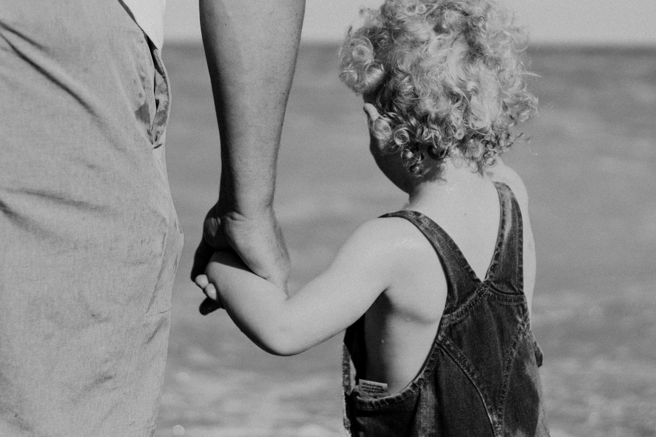 A child with curly hair holding hands with an adult at the beach.