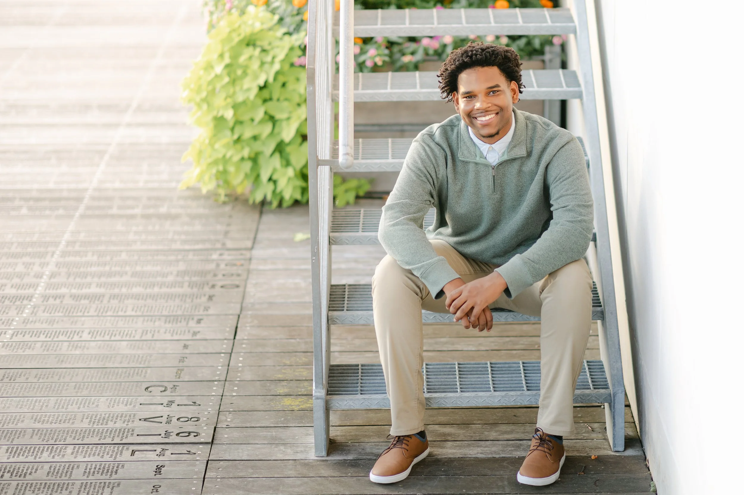 A young man with curly hair, smiling, sitting on outdoor metal stairs, wearing a gray sweater, beige pants, and brown shoes, with greenery and a white wall nearby. Photo taken by Milwaukee, Wisconsin Senior Photographer.
