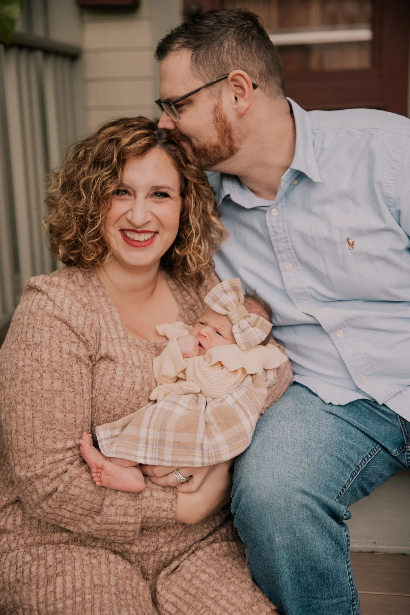 A woman holding a newborn baby, a man leaning in and kissing her on the forehead, they are sitting on front steps outside.