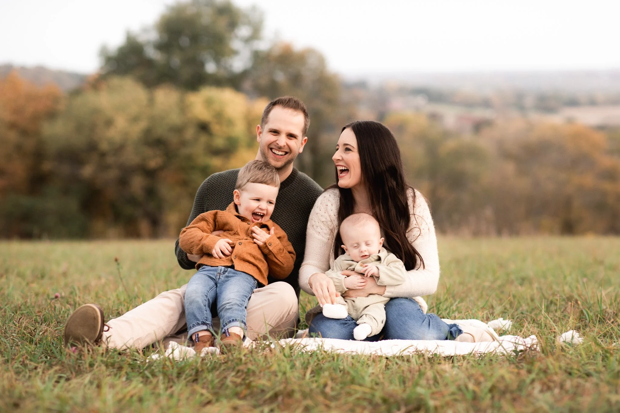 Family of four sitting on a blanket outdoors, smiling and laughing, with fall trees in the background.