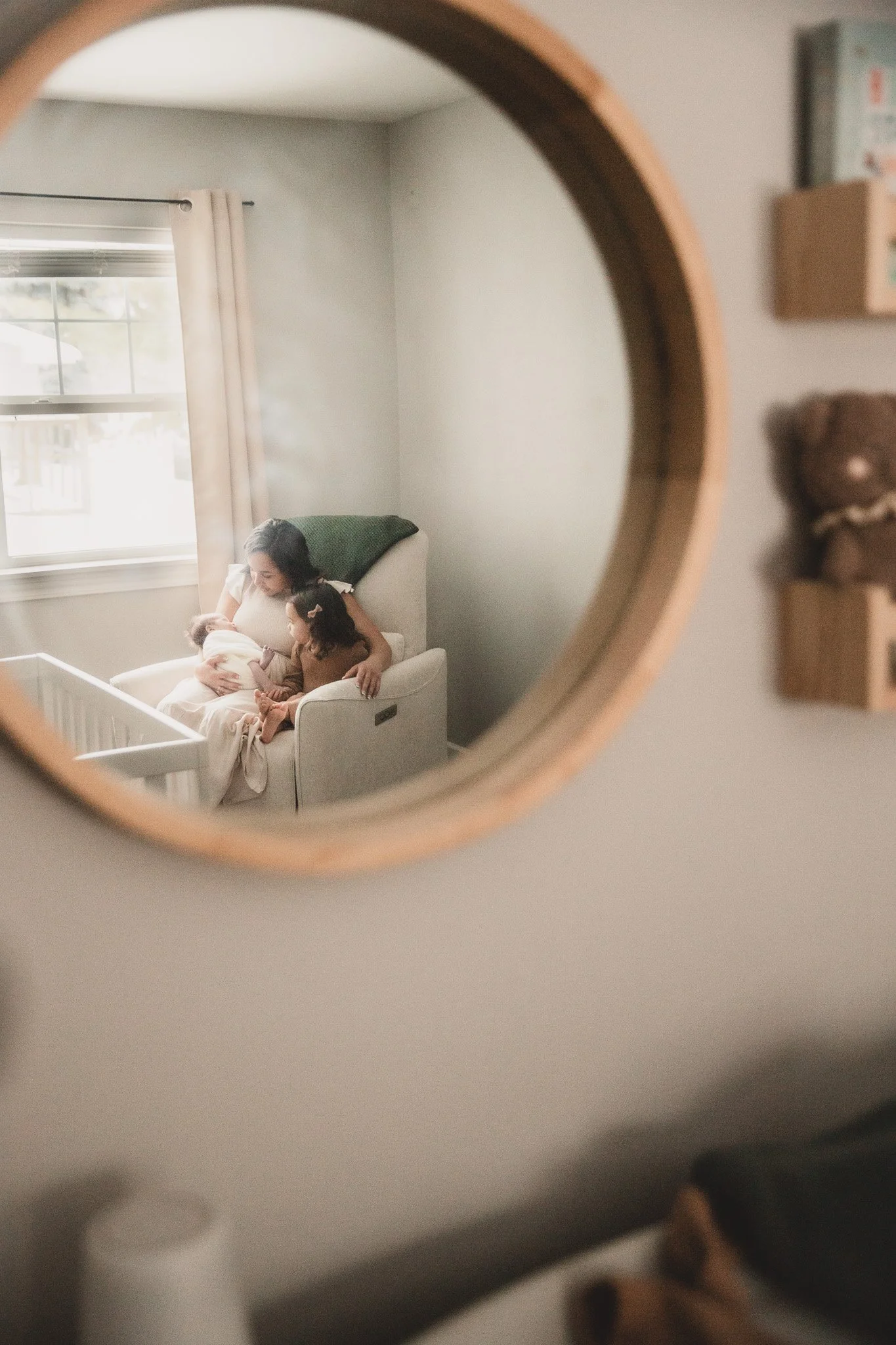 A mother breastfeeding two children while sitting in a nursery, reflected in a round mirror on the wall.