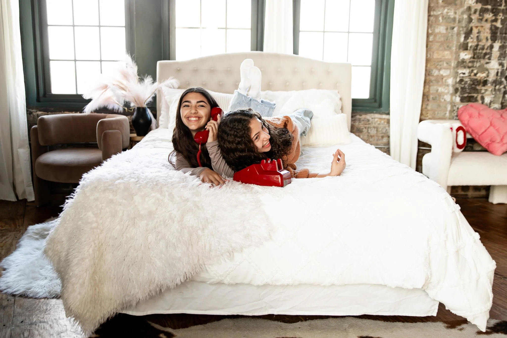 Two young women are lying on a large, white bed with fluffy bedding in a cozy bedroom. One woman is talking on a red vintage rotary phone, smiling, and the other woman is lying next to her, resting her head on the first woman's shoulder, also smiling. The room has large windows, a brick wall, and decorative pillows.