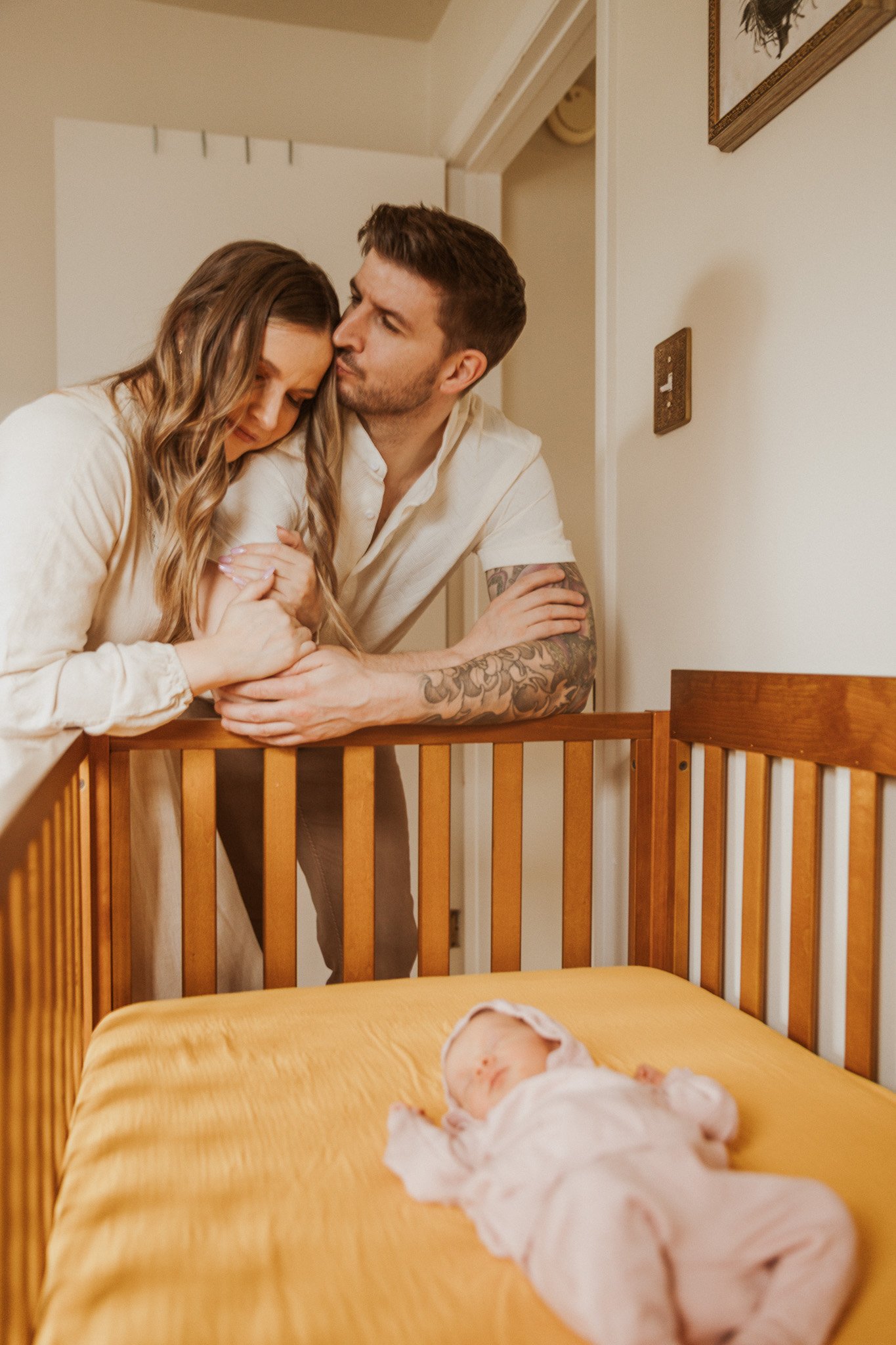 A couple with tattoos leaning on a wooden crib, holding hands, looking at a sleeping baby dressed in pink inside a nursery.