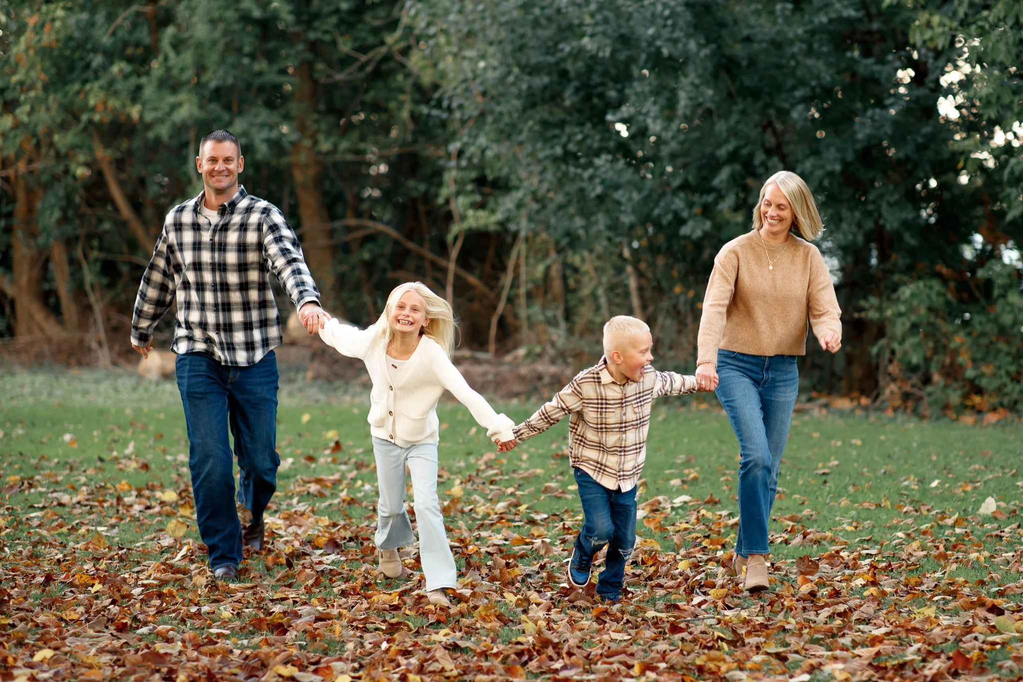 A family of four walking through a park filled with fallen autumn leaves, holding hands and smiling.