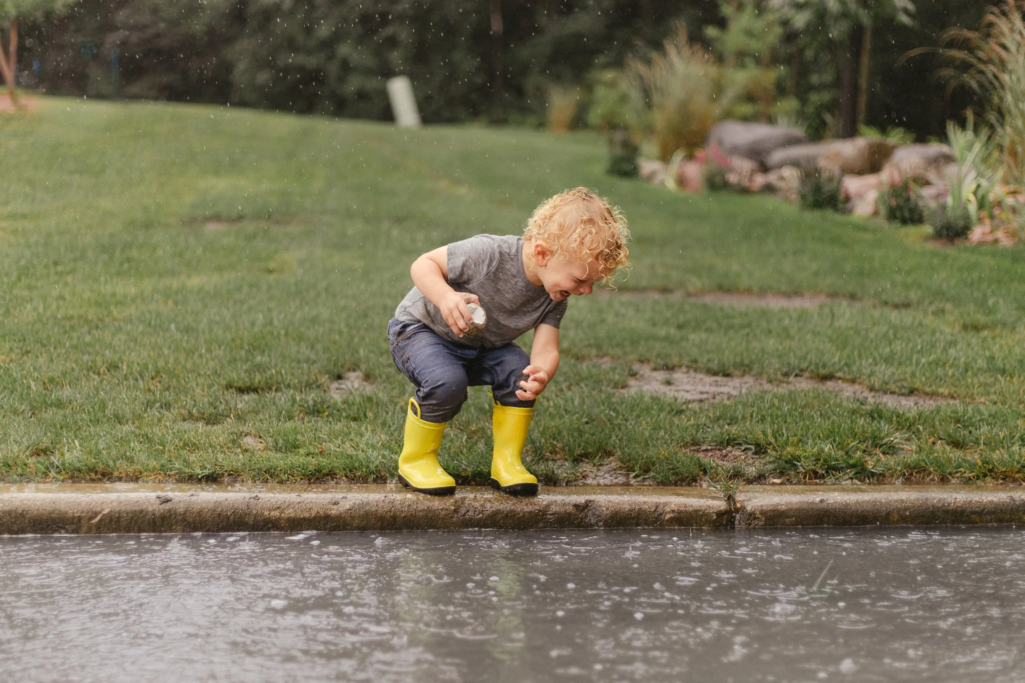A young boy with curly blonde hair wearing a gray t-shirt, dark jeans, and yellow rain boots squatting on the curb by a street, holding a rock in his right hand, looking into a puddle of water. It is raining, and he appears to be playing and enjoying the moment.
