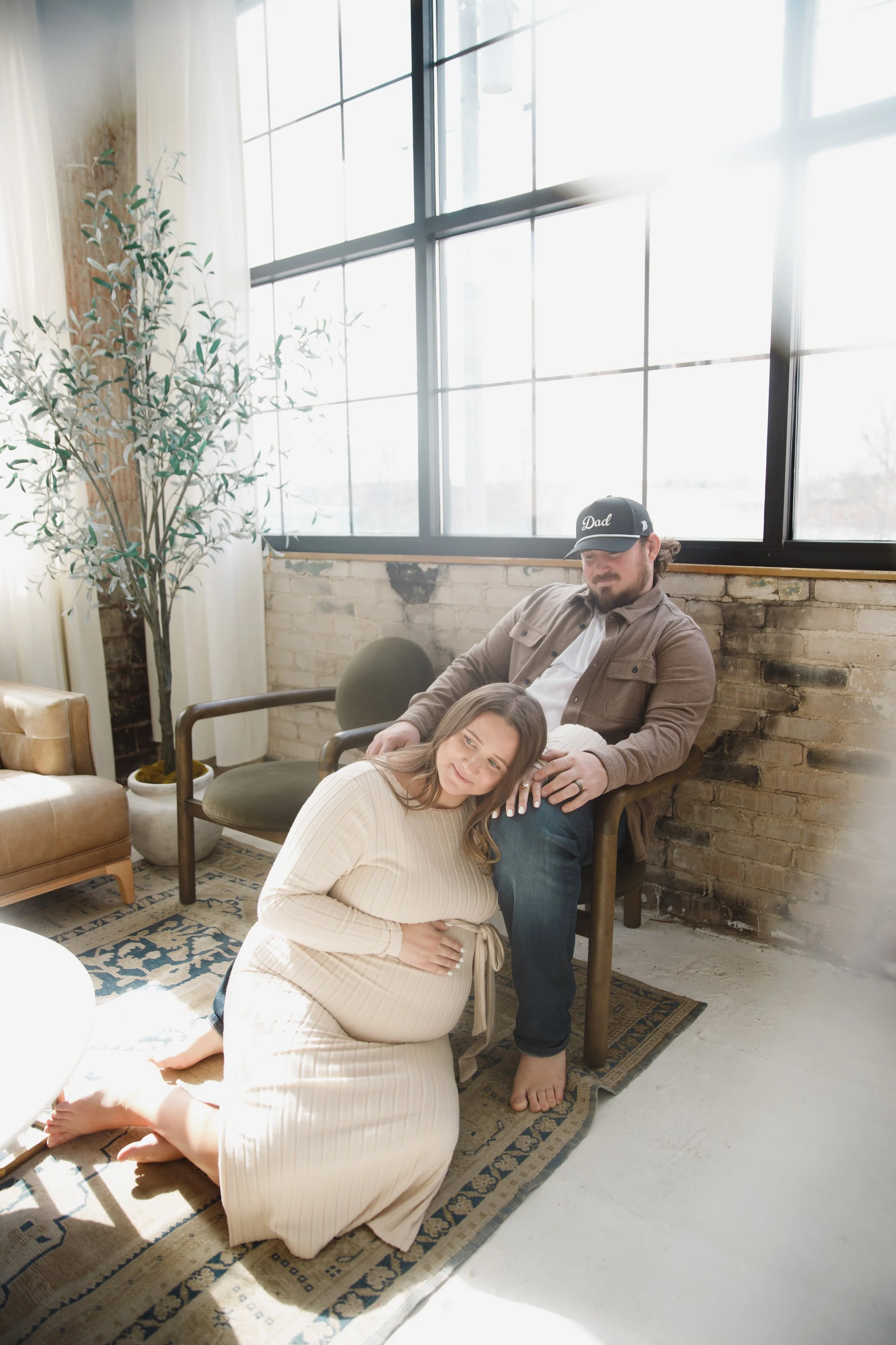 A pregnant woman kneels on a rug with her hand on her belly, leaning on a man sitting in a chair. The man is casually dressed, wearing a cap, in a room with large windows, brick wall, and indoor plants, lit by sunlight. Studio in Milwaukee, Wisconsin