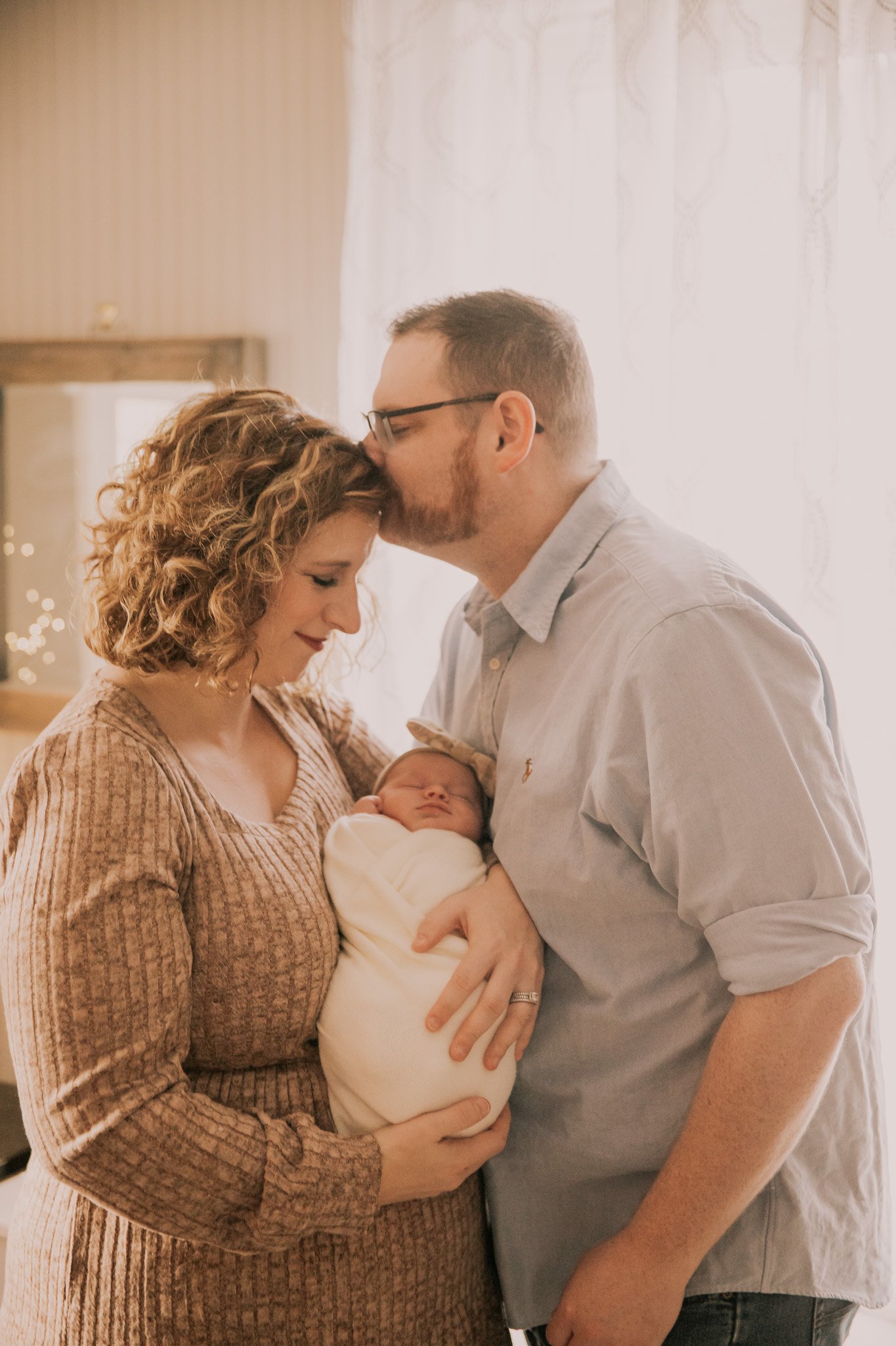 A woman and a man hold a newborn baby wrapped in a blanket. The man is kissing the woman's forehead, and they are standing in a softly lit room.
