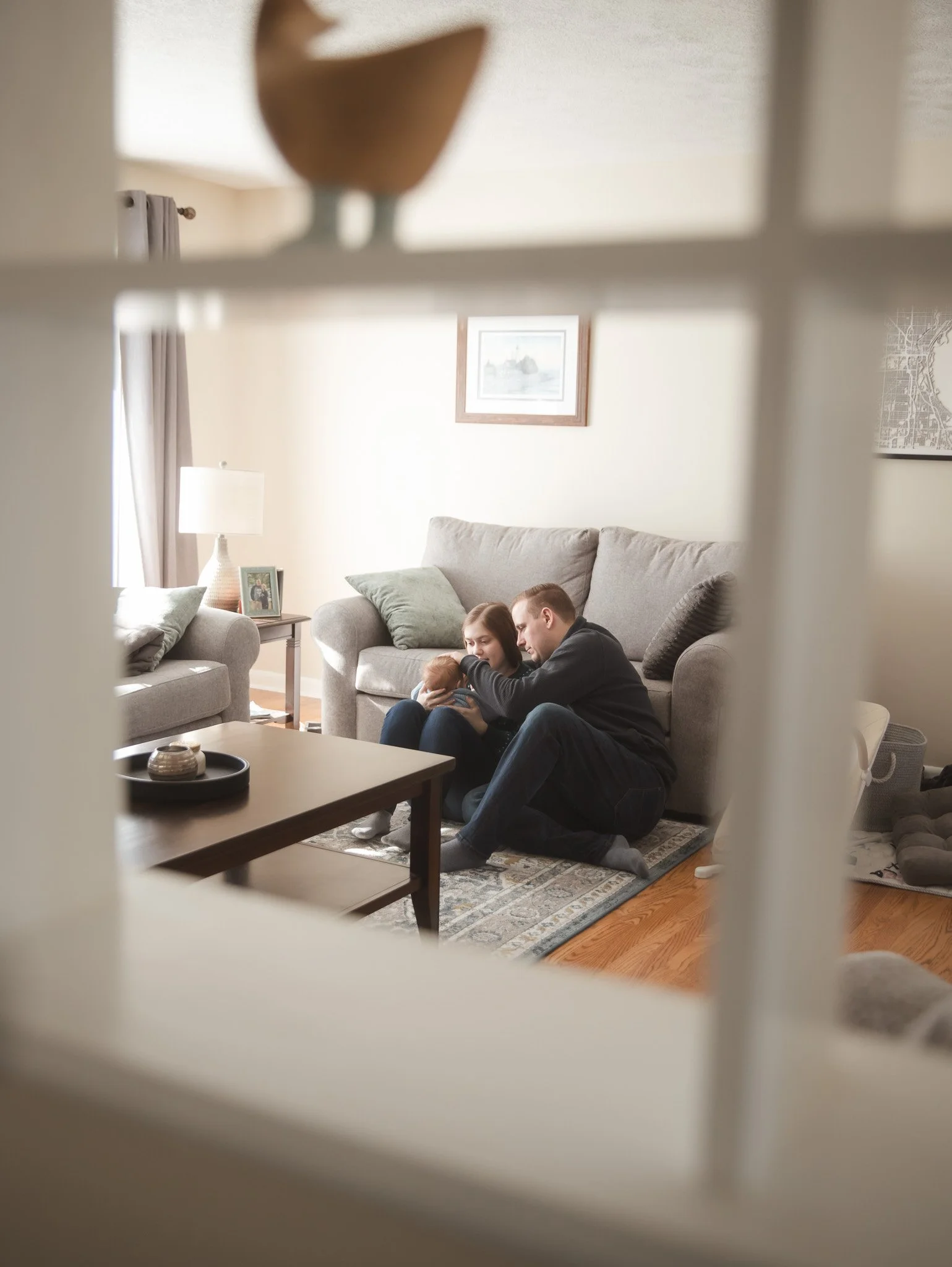 A family sitting on a rug in front of a gray sofa, cuddling and looking at a smartphone, in a cozy living room with framed pictures and a lamp.