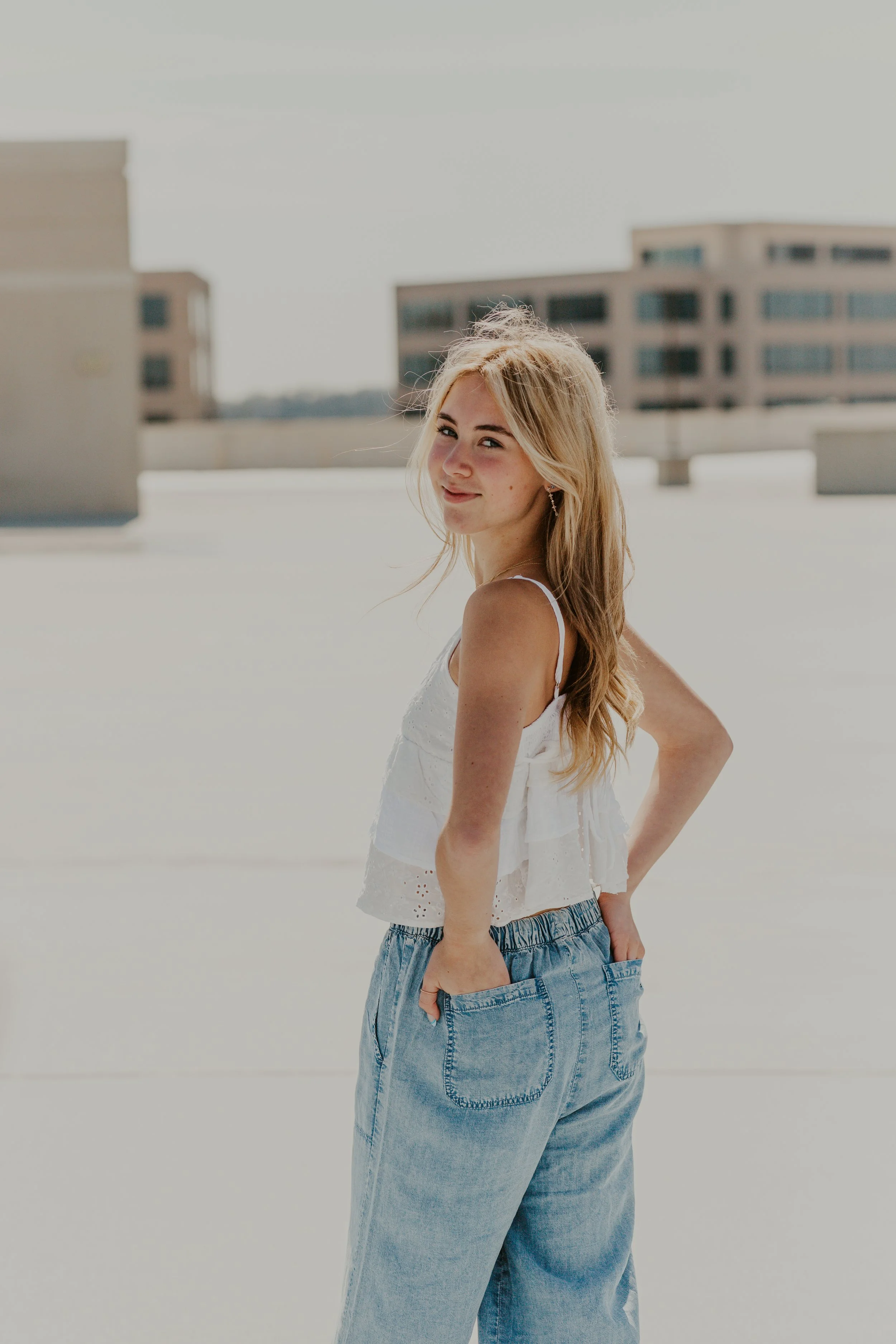 A young woman with long blonde hair in a white sleeveless top and light blue jeans stands on a rooftop during daytime.