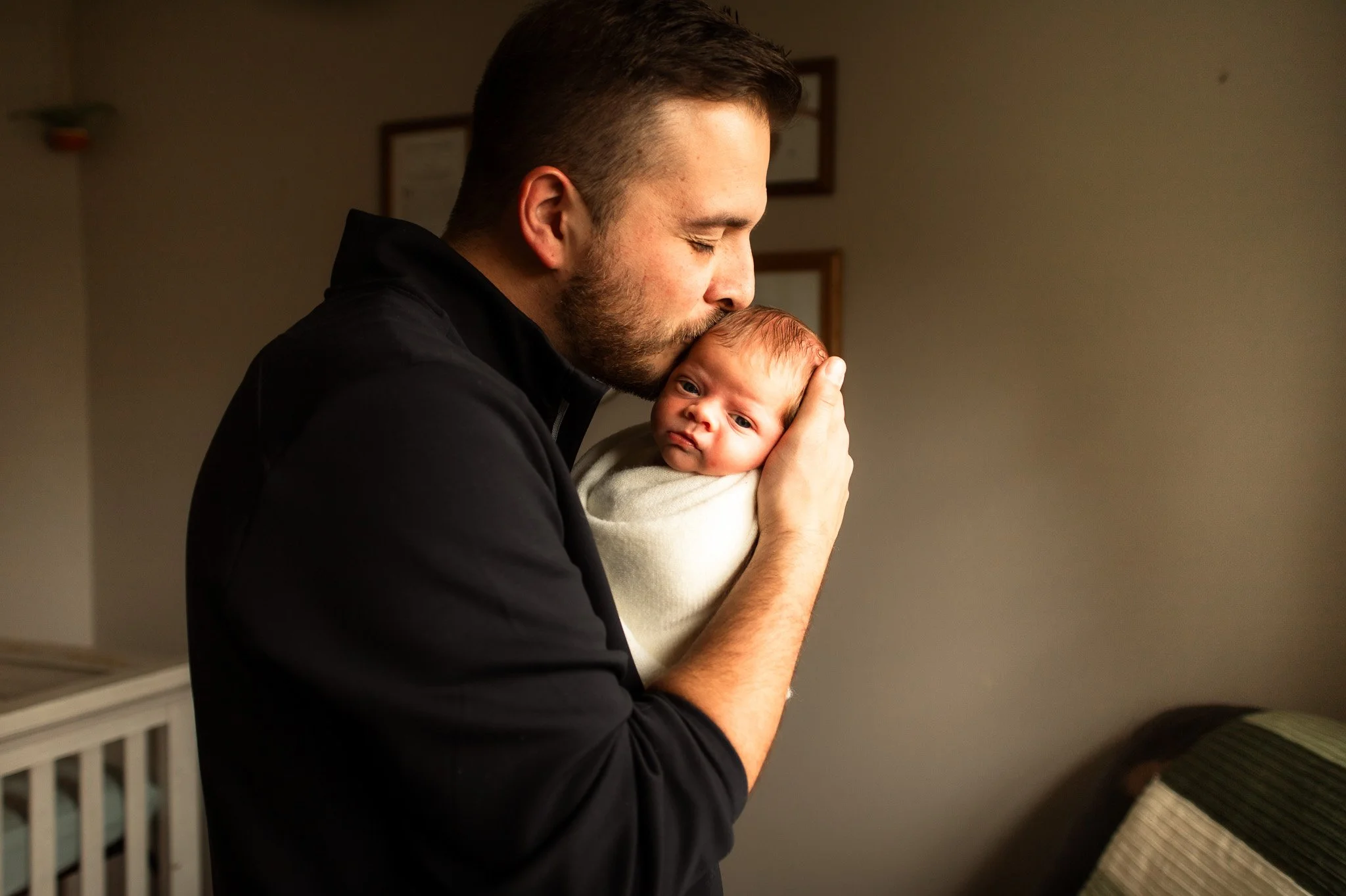 A man holding a wrapped baby close, given a gentle kiss on the head.