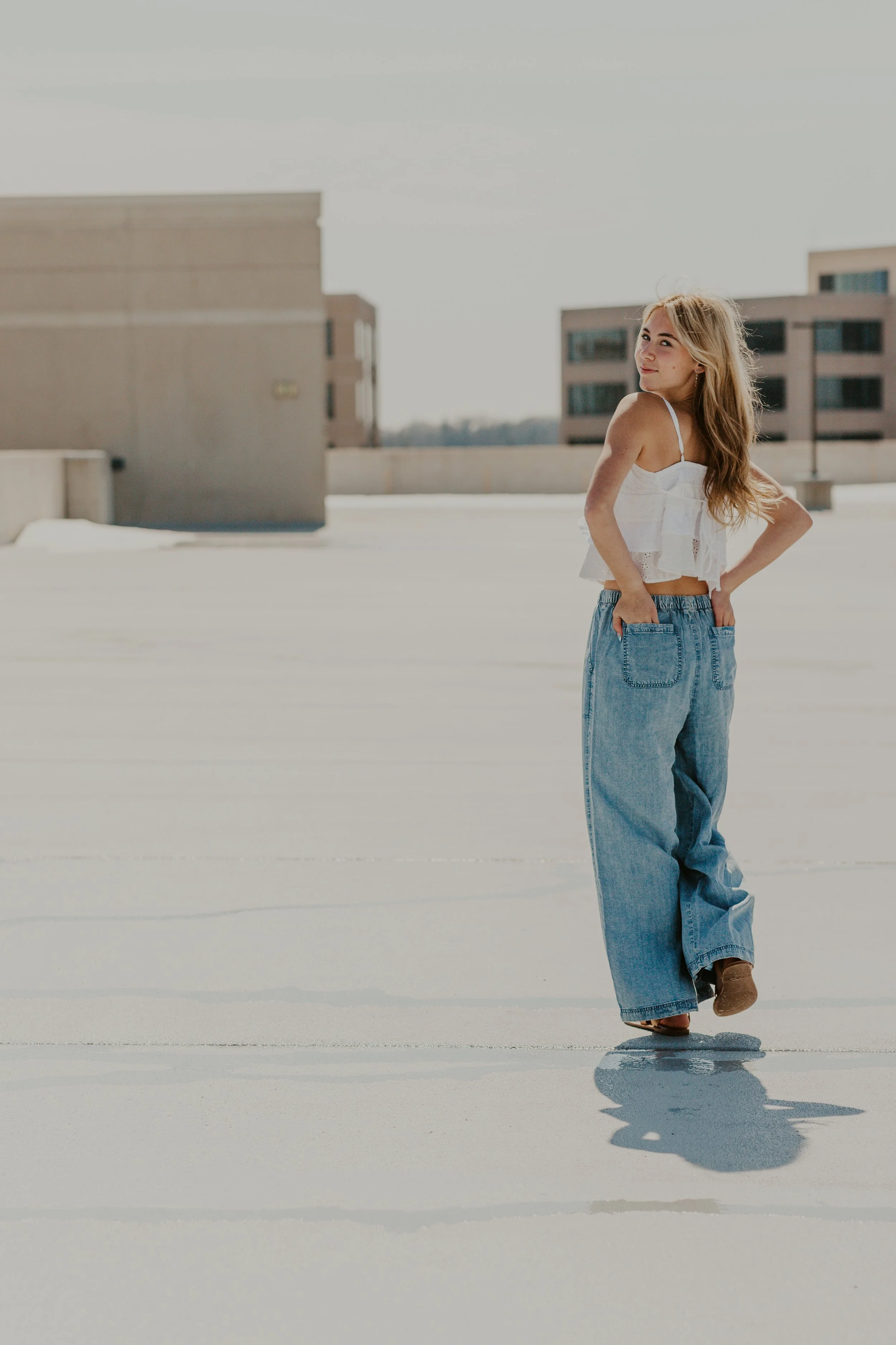 A young woman with long wavy hair stands on a rooftop, wearing a white crop top and loose-fitting blue jeans, with her hands in her pockets, looking back over her shoulder. Photographer in Franklin, Wisconsin.