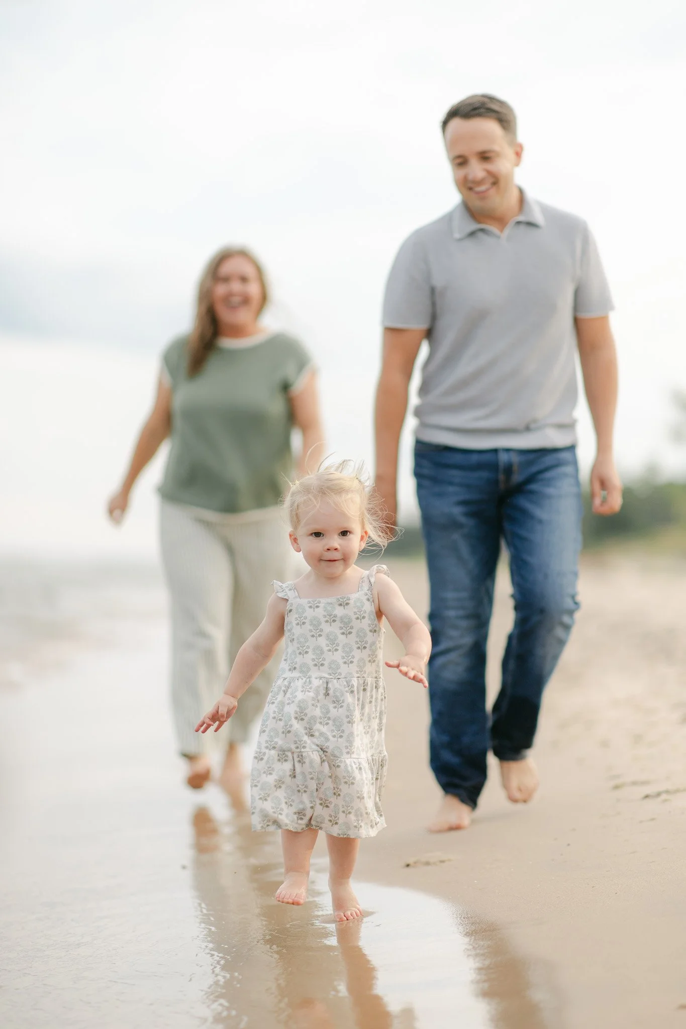 A family walking barefoot on the beach, with a young girl in the foreground and two adults and another person in the background.
