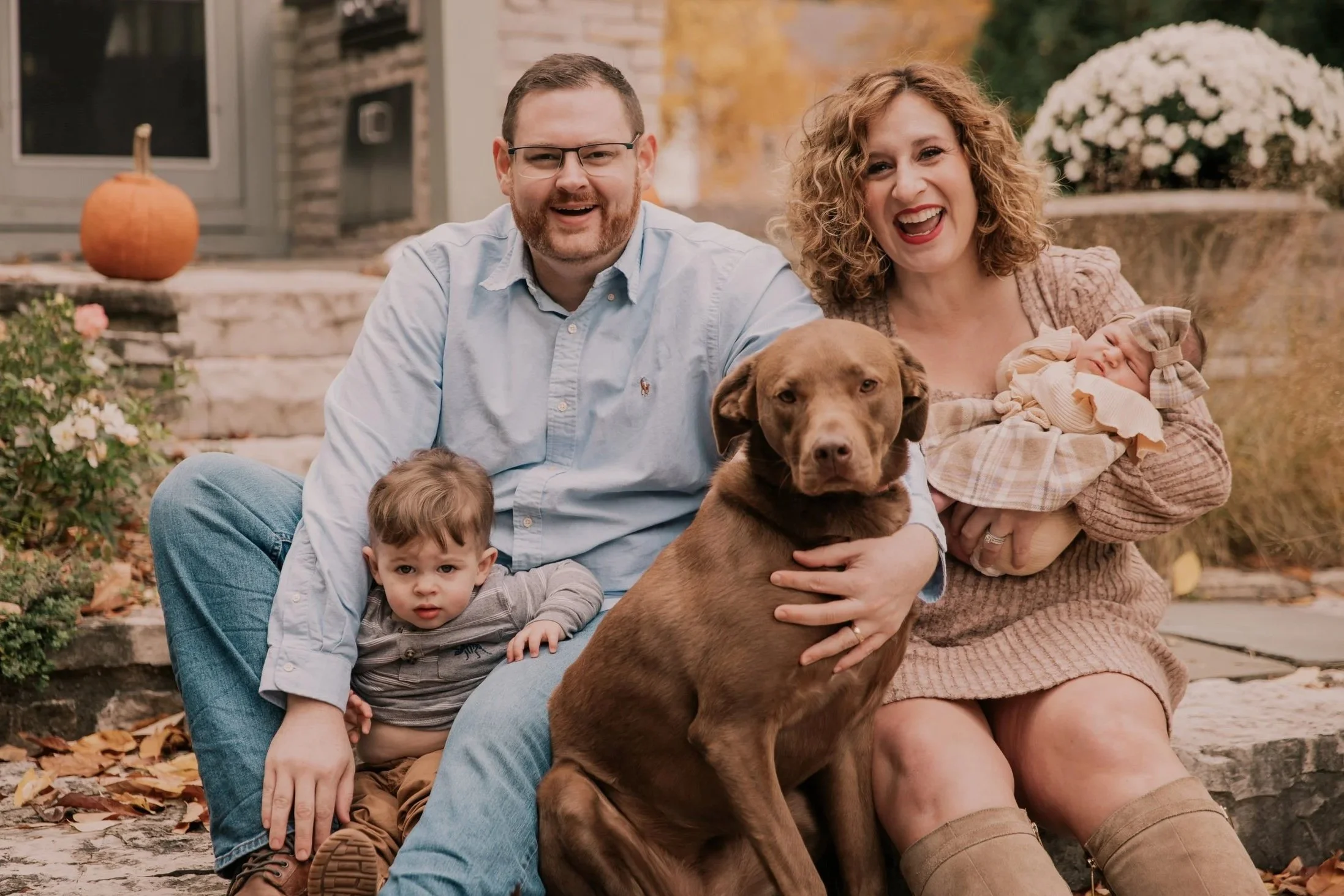 Family of four with a dog, sitting outdoors in fall. The father, with glasses and a beard, is smiling; the mother, with curly hair, is holding a baby girl, and their young son is sitting next to them on the ground. The family is sitting on stone steps with autumn leaves and pumpkins around, and a bush with pink flowers in the background.