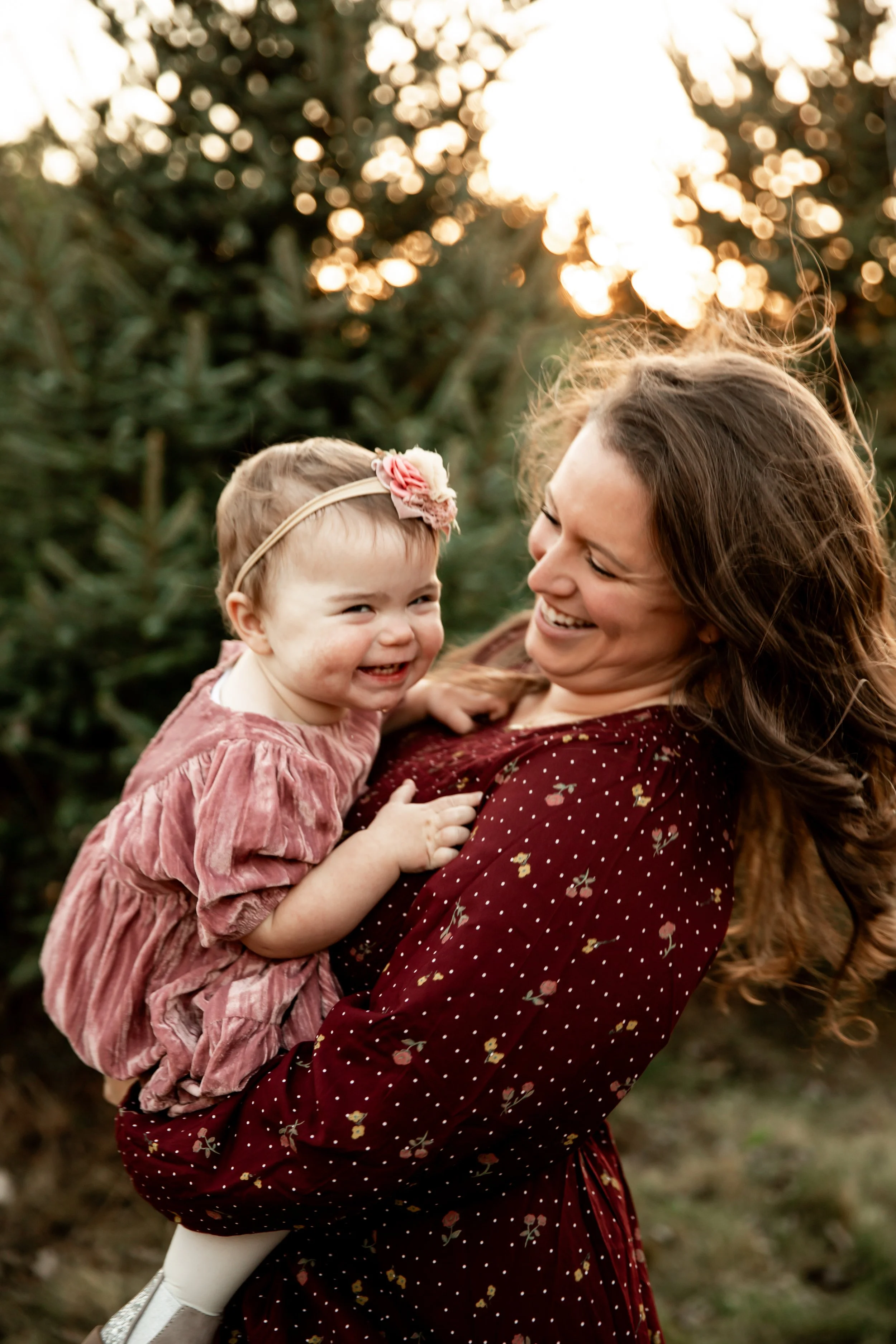 A woman holding a laughing young girl in an outdoor setting during sunset, with trees in the background.
