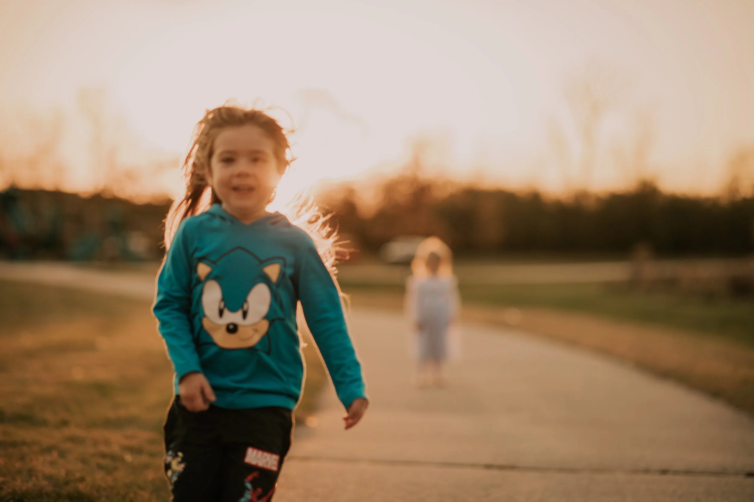 Young girl running outdoors on a sidewalk during sunset, wearing a blue Sonic the Hedgehog hoodie, with a blurred person in the background.