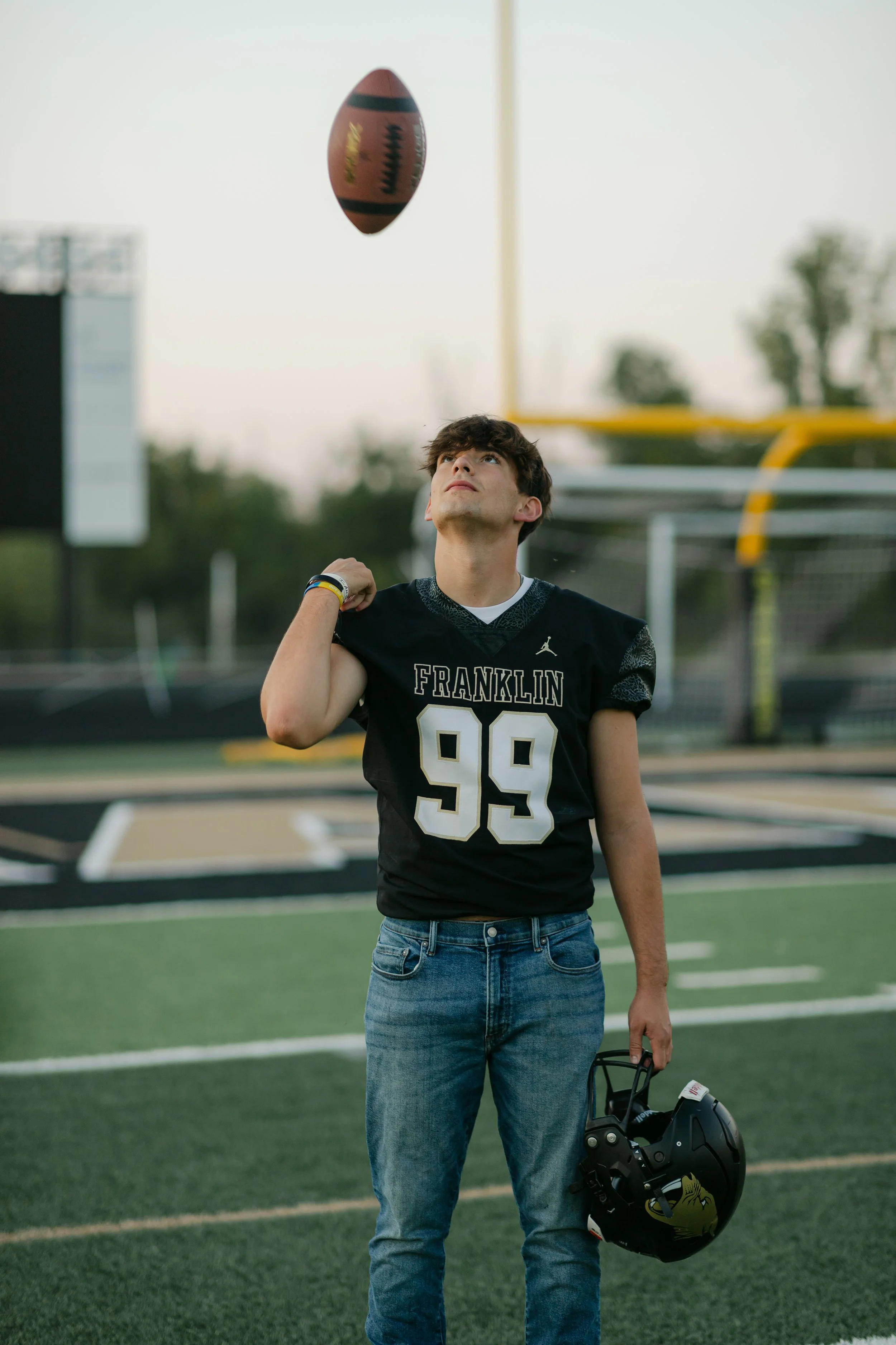 A young football player in a black jersey with the number 99 and the word 'Franklin' on it, standing on a football field and looking up as a football floats above him. Photo taken by Franklin, Wisconsin Senior Photographer.