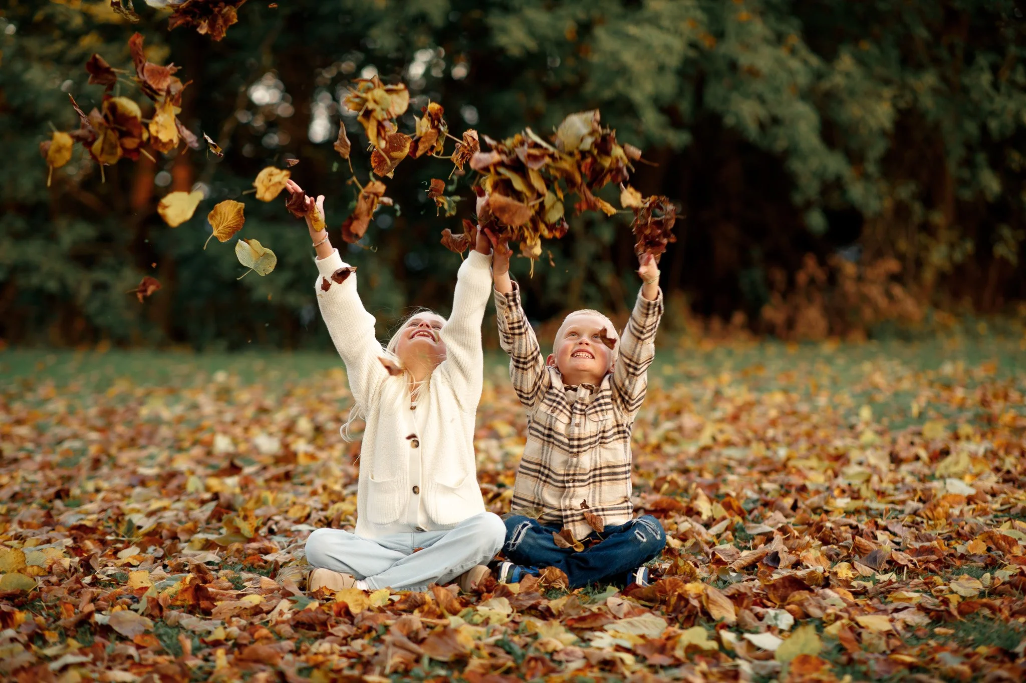 Two children, a girl and a boy, sitting on a blanket in a park during autumn, playing with fallen leaves and throwing them into the air, smiling and enjoying the season.