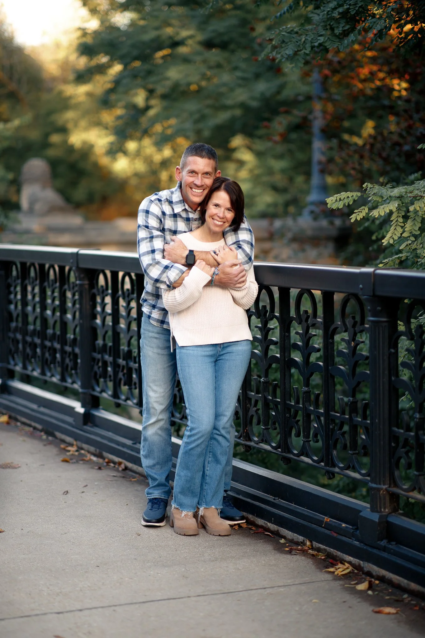 A smiling couple standing on a sidewalk near a black decorative iron fence in a park with green trees and autumn leaves.