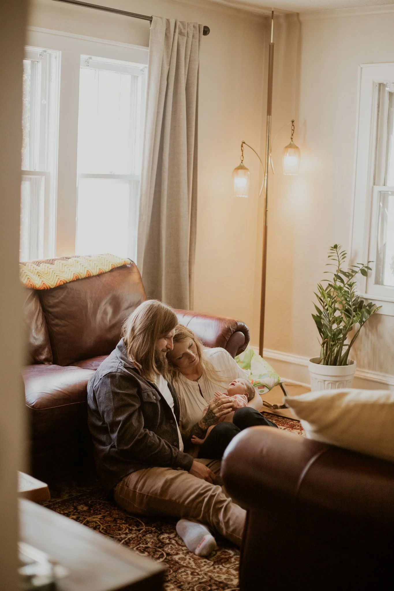 A family sitting on the floor of a living room, with parents and a newborn baby, near a brown leather couch, lit by natural light coming through windows with beige curtains.