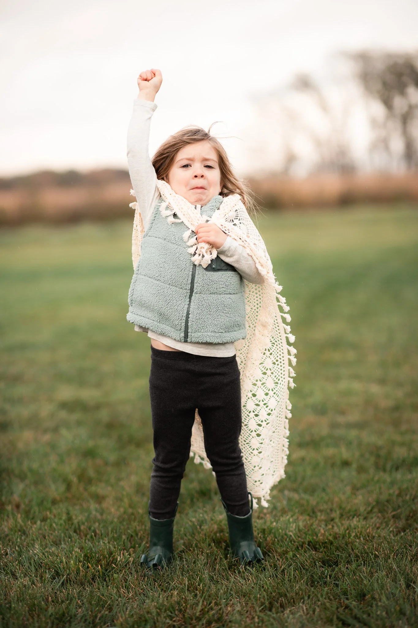 A young girl standing outdoors on grass, raising her right fist in the air with a determined expression. She is wearing a gray fleece vest, black pants, green rain boots, and has a cream-colored crocheted blanket draped over her shoulders.