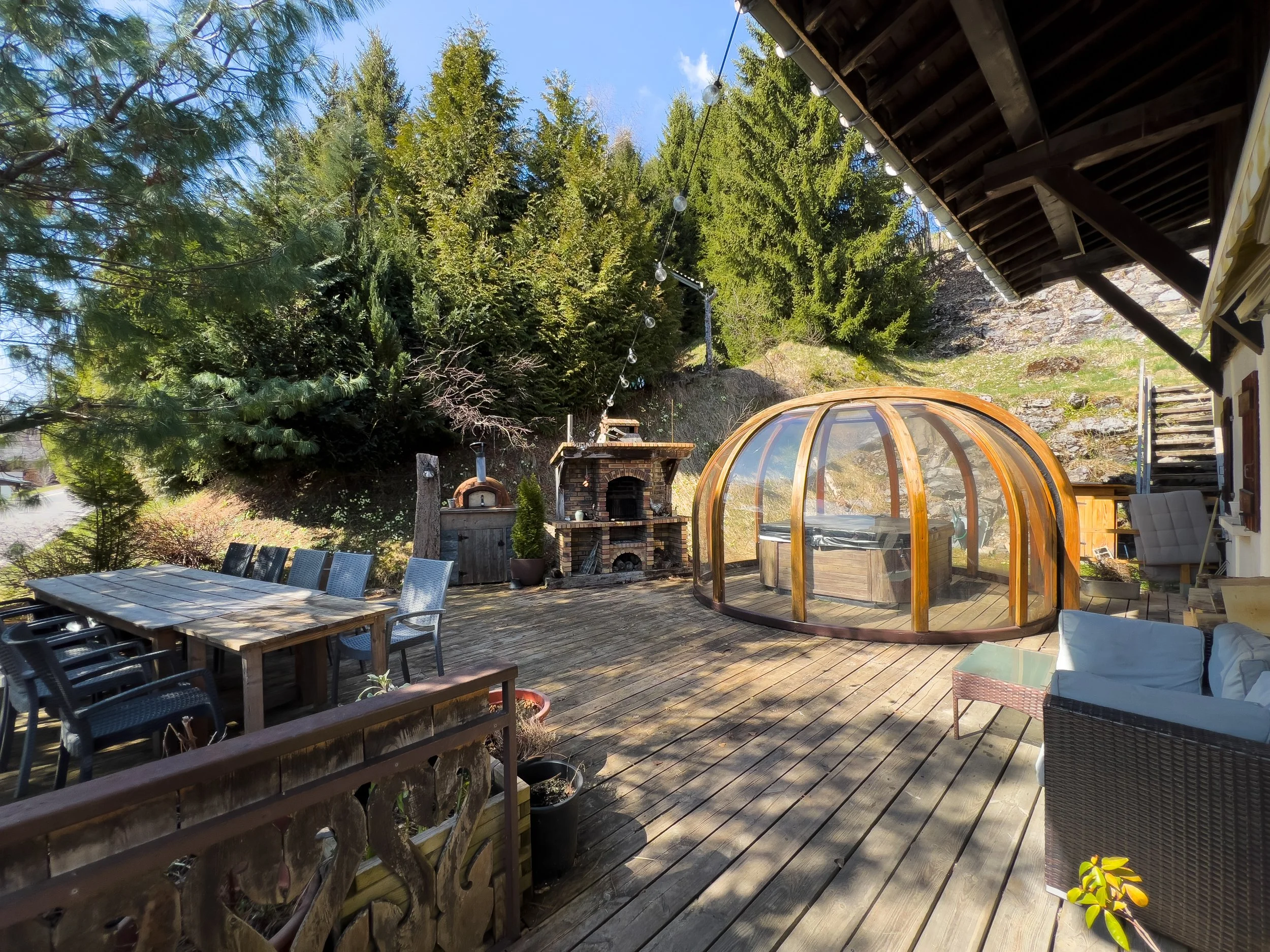 Outdoor terrace at Chalet Sandy in Les Gets with large dining table, wood-fired pizza oven, and Alpine forest setting.