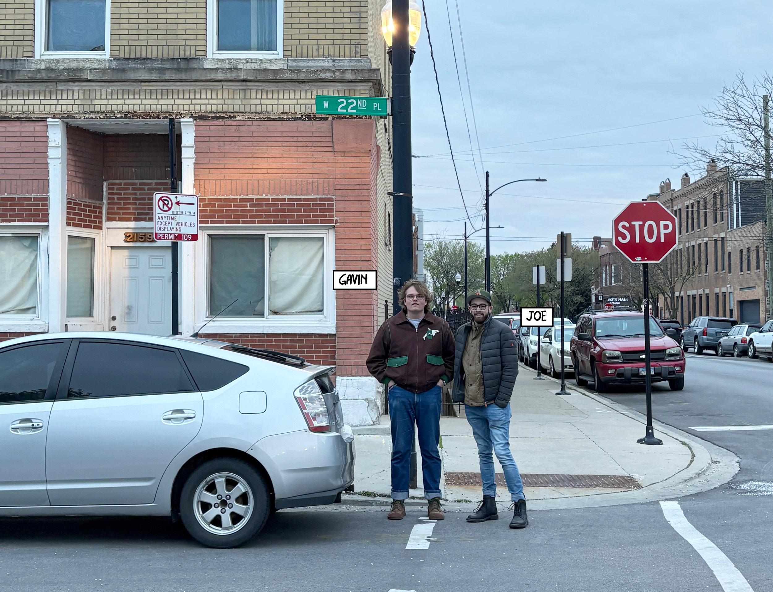 Two young men, identified as Gavin and Joe, are standing on a Chicago sidewalk at an intersection near a stop sign, with parked cars and buildings in the background, during the day.