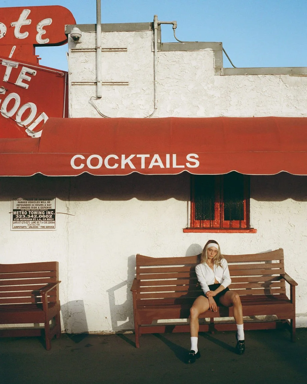 A young woman sitting on a wooden bench outside a bar or restaurant with a red awning labeled 'COCKTAILS'. There are two benches, she is wearing a white button-up shirt, black shorts, white socks, and black shoes, with light-colored hair and a headba