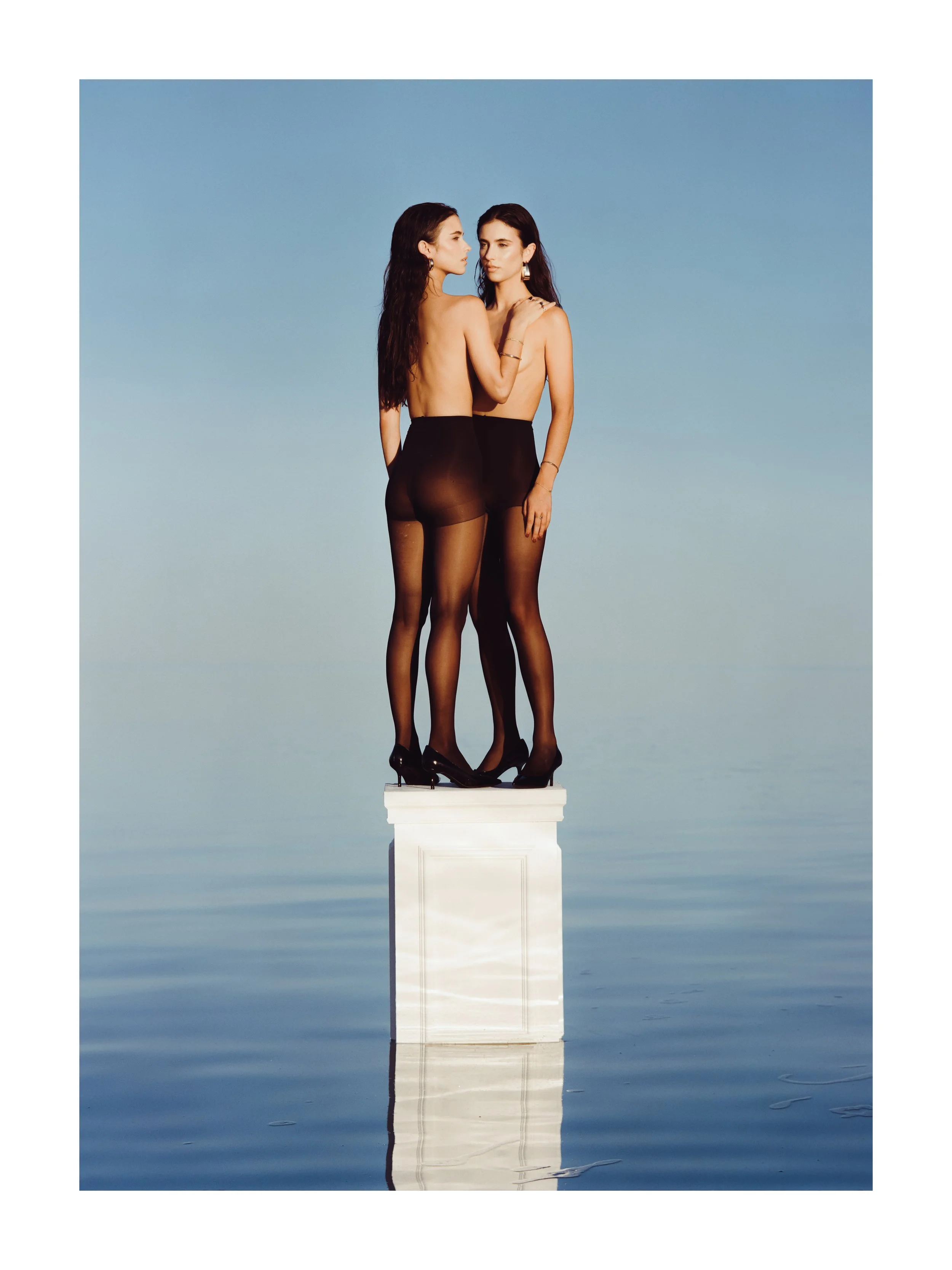 Two women with long dark hair standing close together on a white pedestal, wearing black sheer stockings and high heels, against a blue sky background.