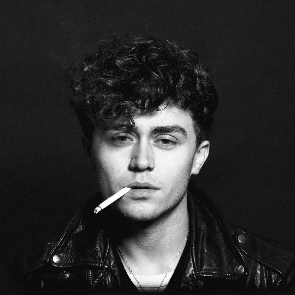 Black and white portrait of a young man with curly hair, wearing a leather jacket, smoking a cigarette.