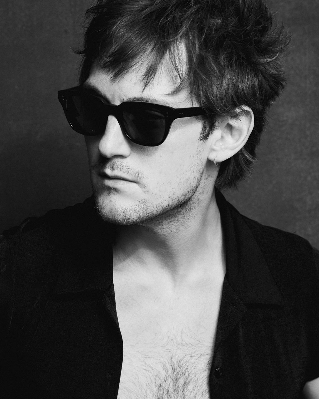 A young man with messy hair, wearing dark sunglasses and a partially unbuttoned black shirt, looking to the side in a black-and-white portrait.
