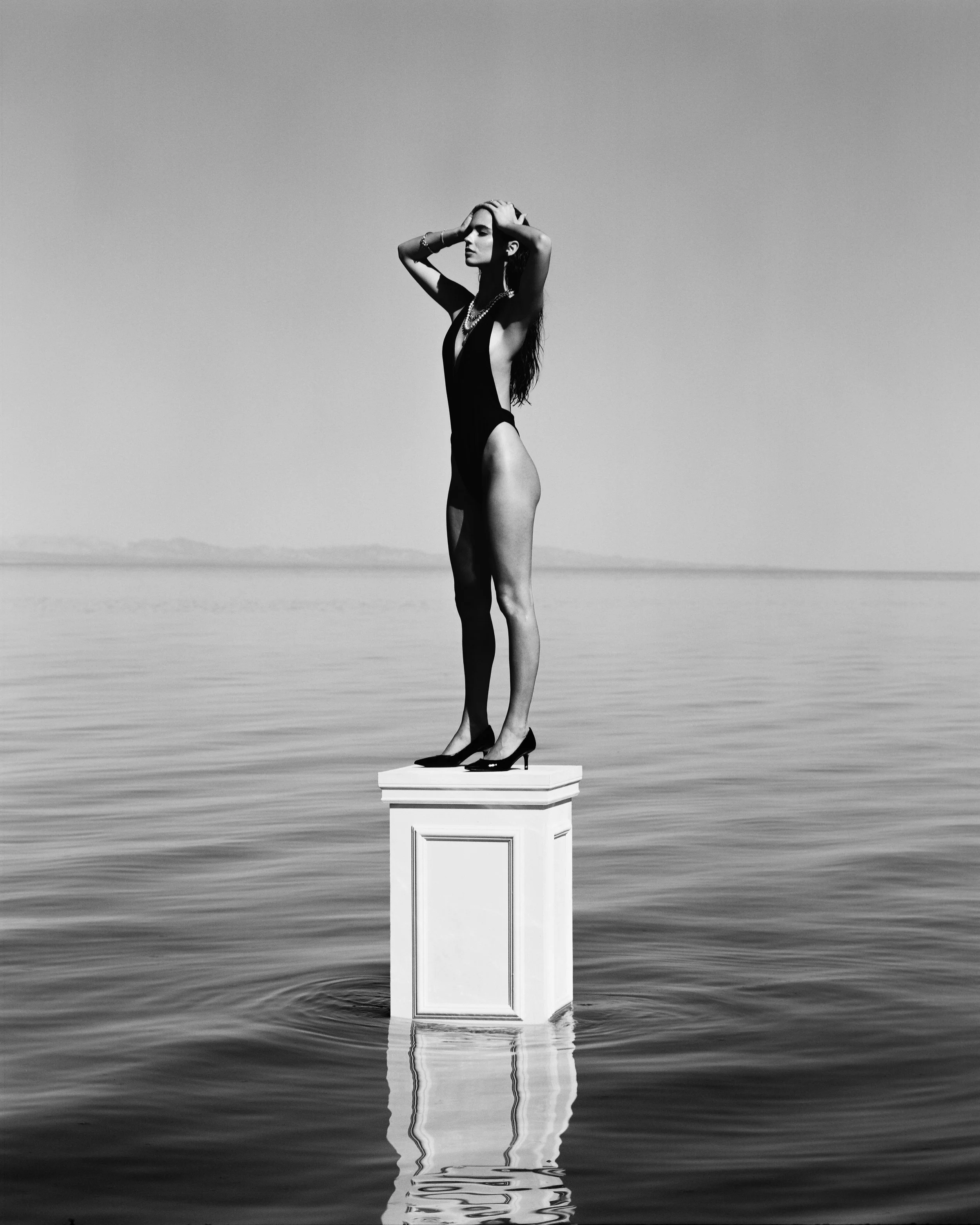 A woman in a black swimsuit and high heels standing on a pedestal in the water, with her hands on her head, in a black and white photo.