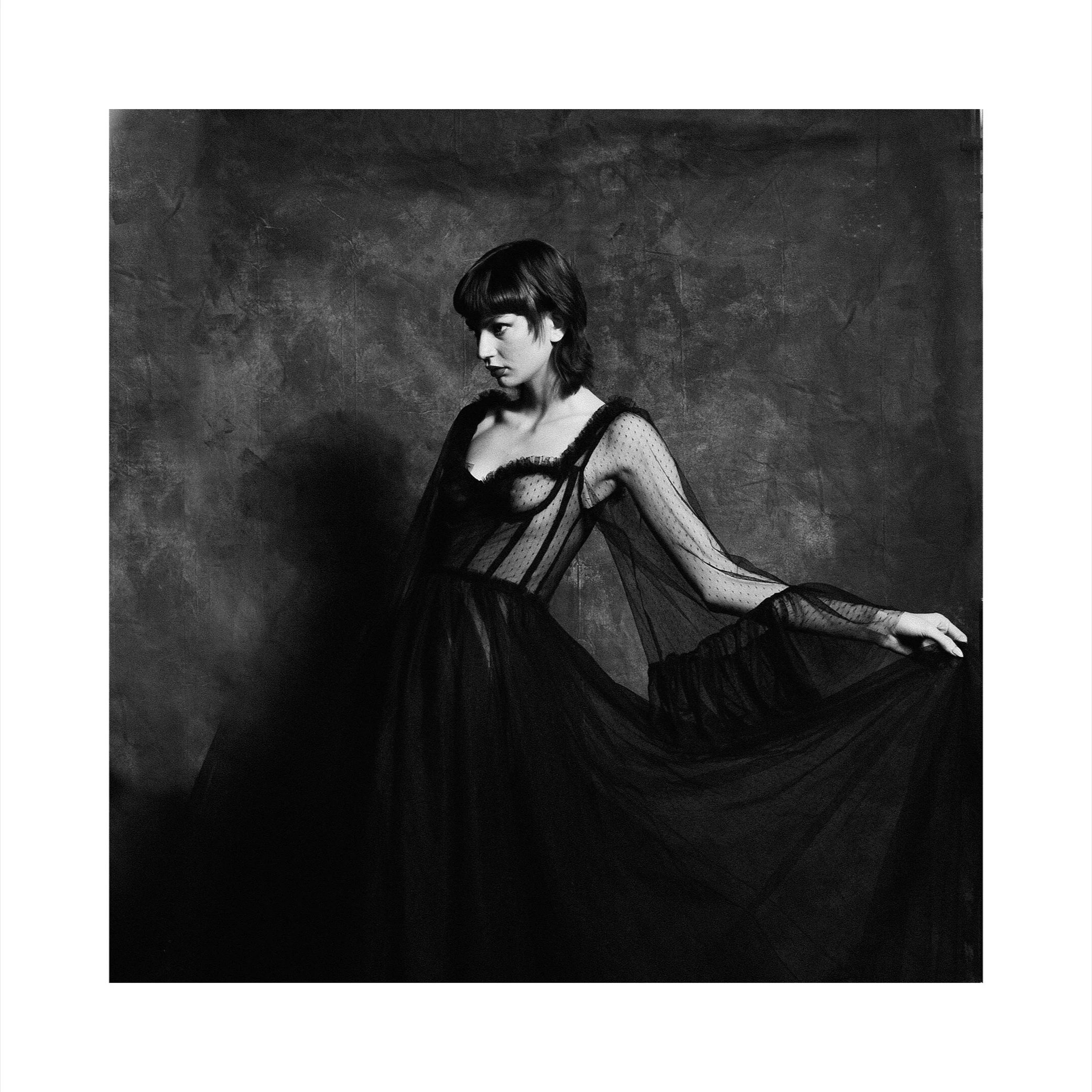 Black and white photo of a woman wearing a sheer, polka-dot dress with ruffled details, posed against a textured background.