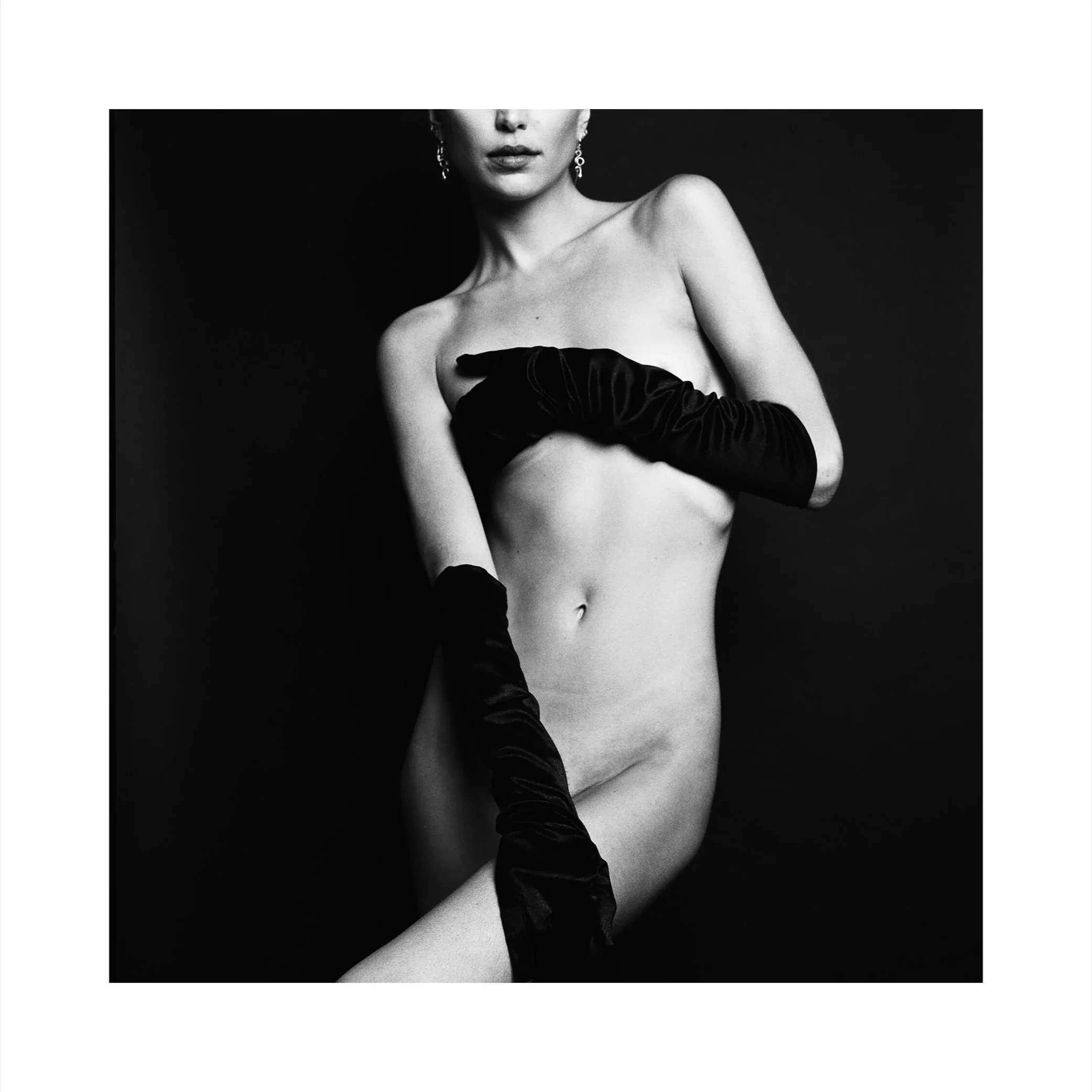 Black and white photo of a topless woman wearing long black gloves, posing against a dark background.