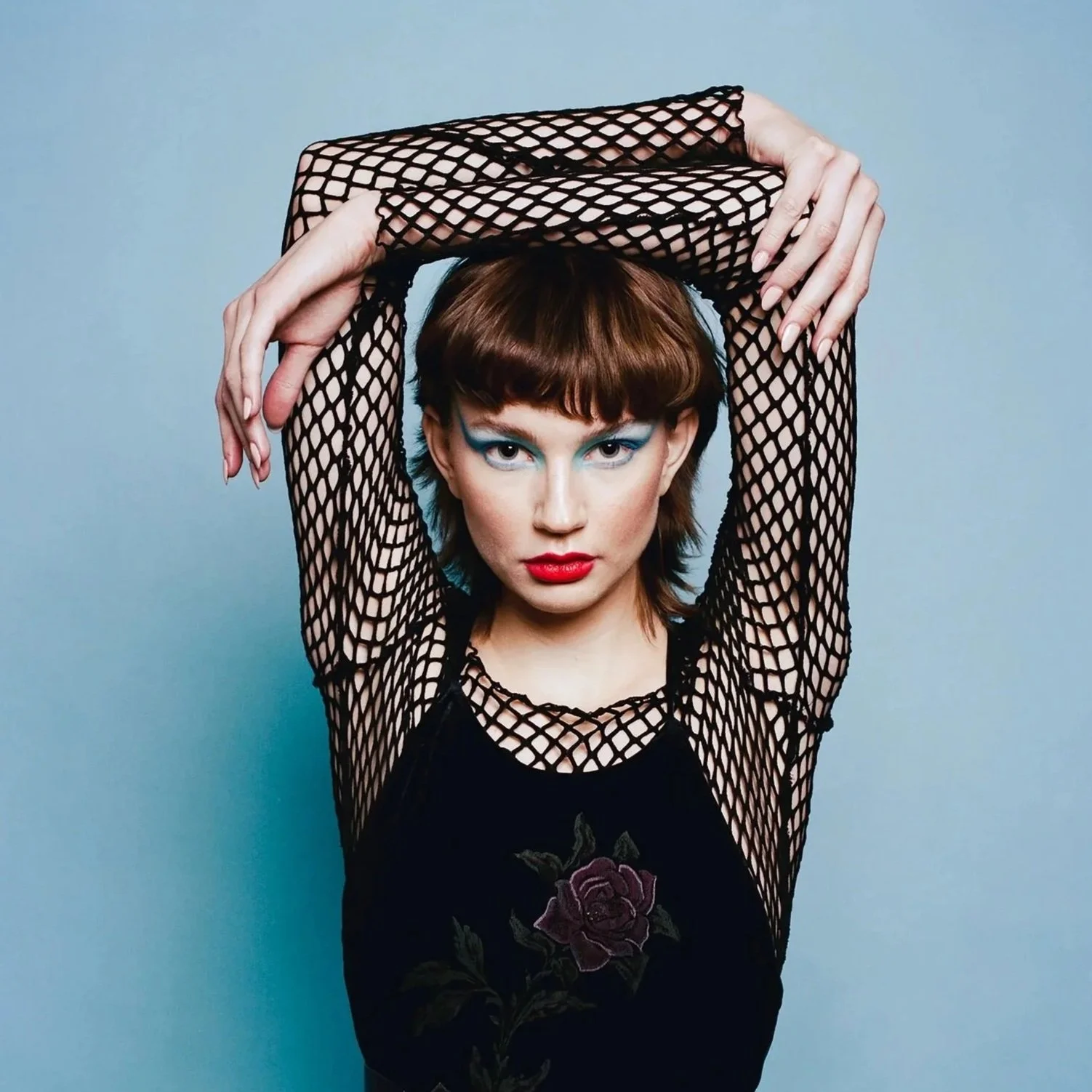 A woman with short brown hair and bold makeup poses with her arms raised above her head, wearing a black fishnet top and a black velvet dress with a rose embroidery.