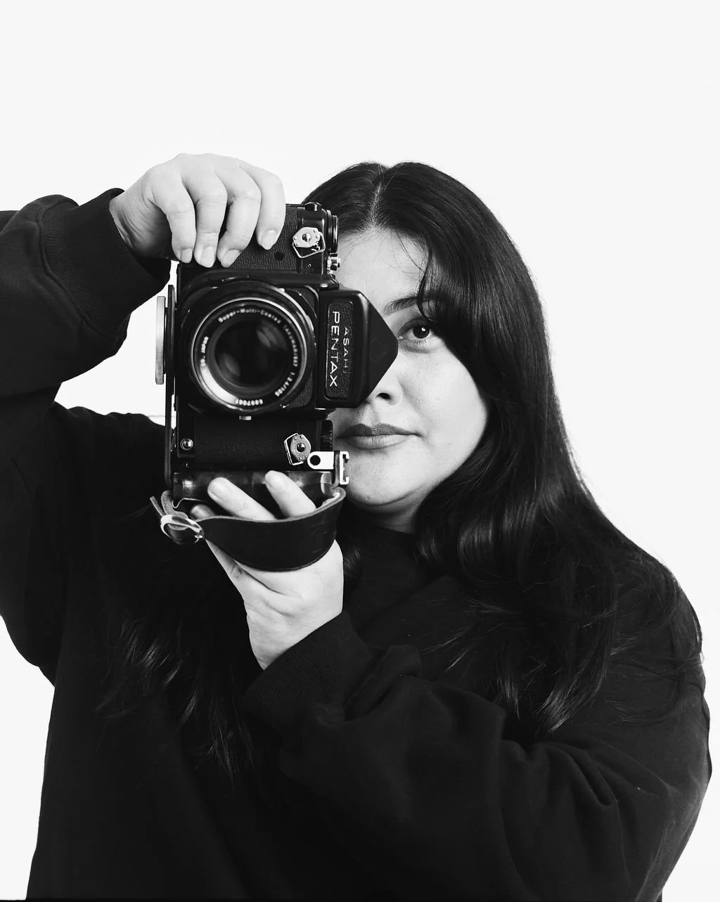 Woman with long dark hair taking a black-and-white photograph with a Pentax camera.