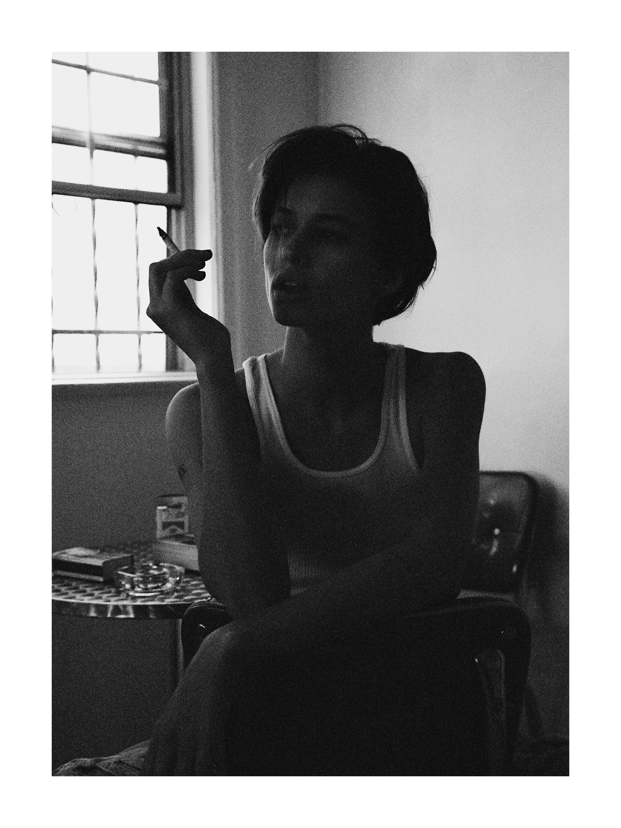 A woman sitting indoors near a window with bars, holding a cigarette near her face, shadows cast across her features in a black-and-white photograph.