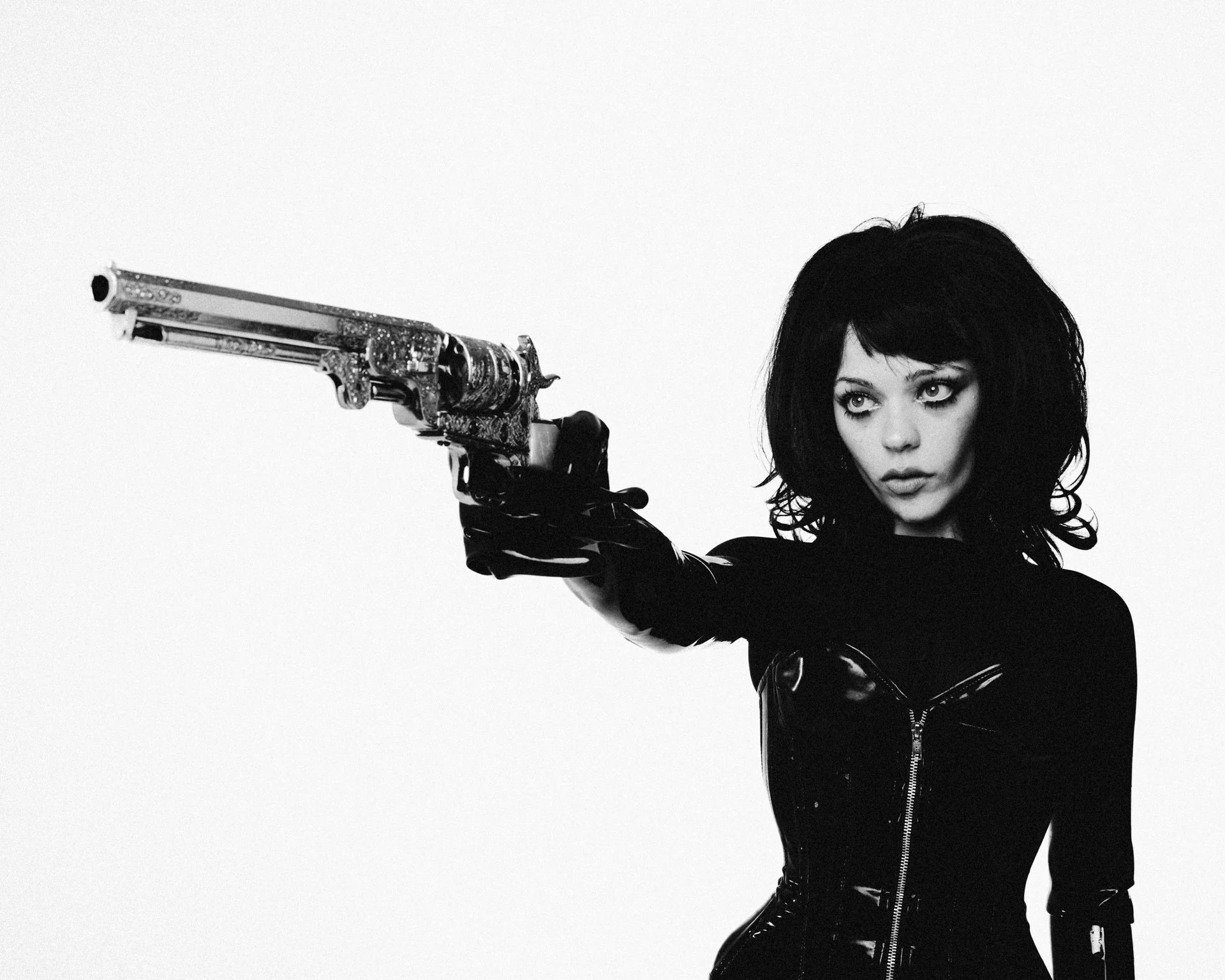 A woman with dark, voluminous hair and intense eyes, wearing a black leather jacket, points a gun directly at the camera.