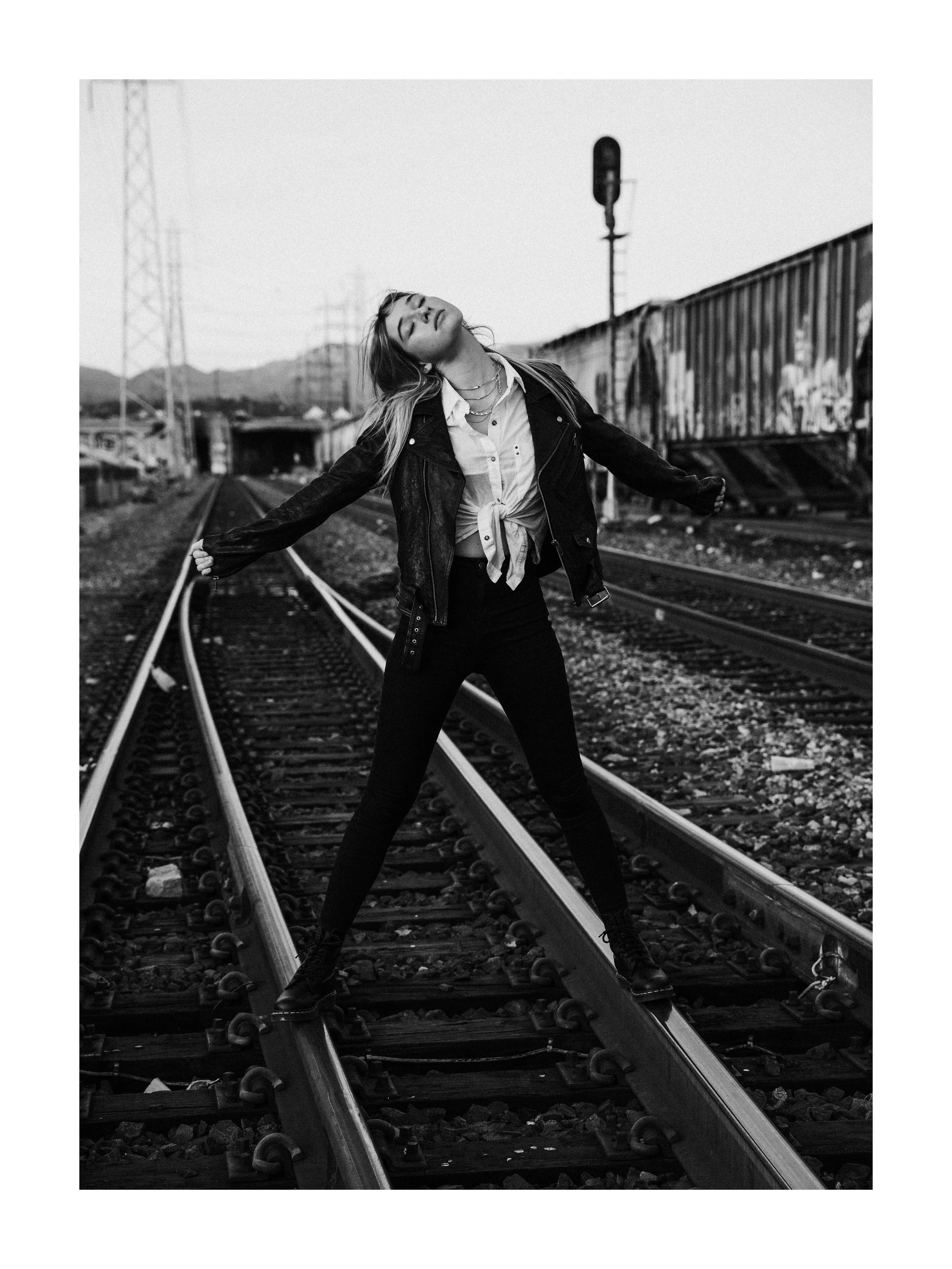 A woman standing on train tracks with arms outstretched and head tilted back in a relaxed pose, wearing a leather jacket and tied shirt, with train cars in the background.