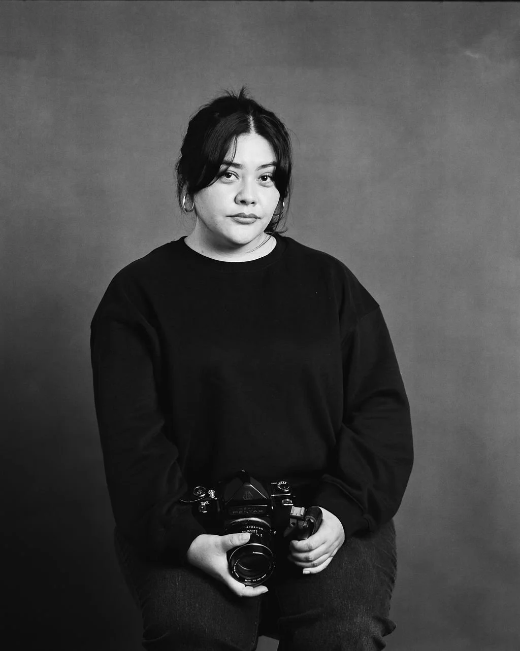 A young woman with dark hair, hoop earrings, and wearing a black sweatshirt holds a camera in her hands. She is sitting against a plain background.