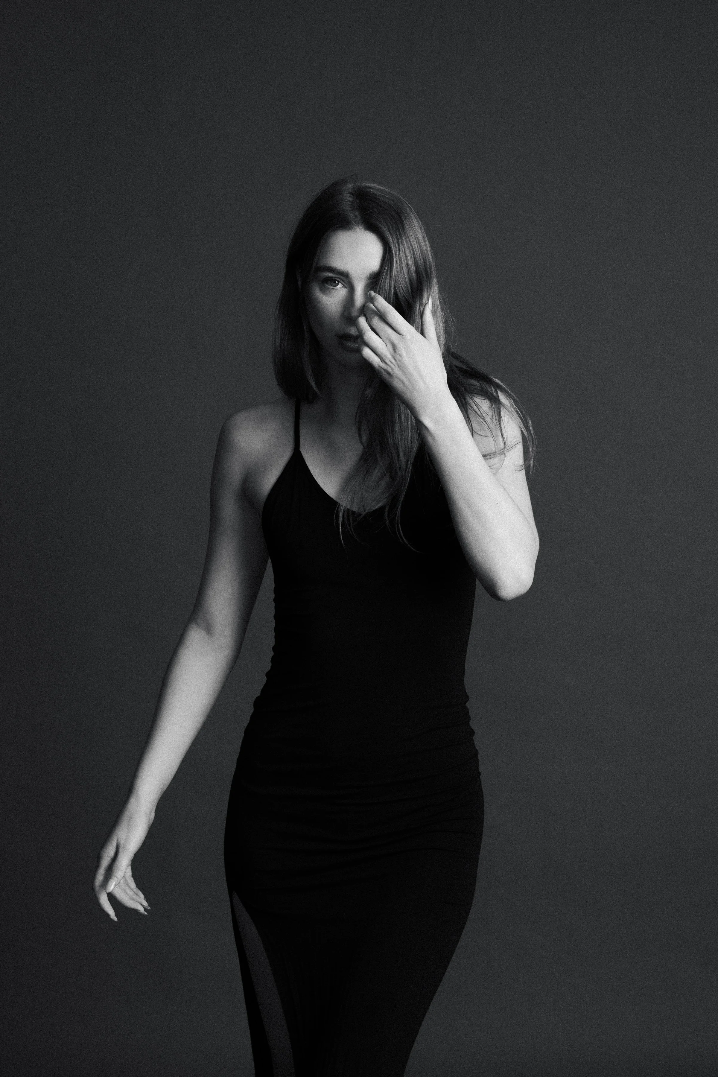 Black and white photo of a woman with long hair wearing a sleeveless black dress, touching her face with her left hand, against a dark background.