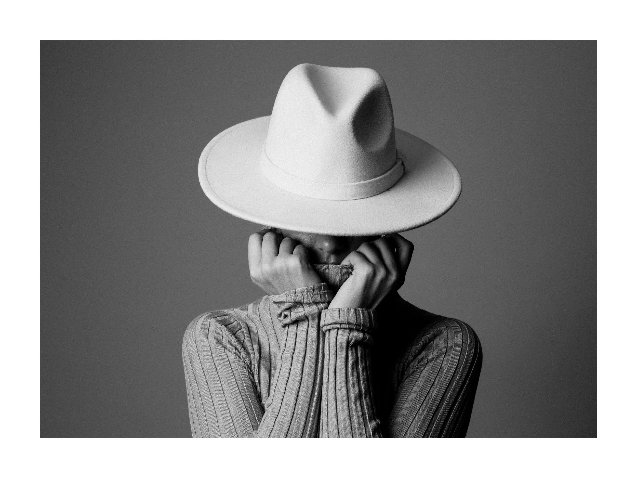 Person wearing a large white hat and a striped long-sleeve shirt, covering their face with their hands in a black and white photo.